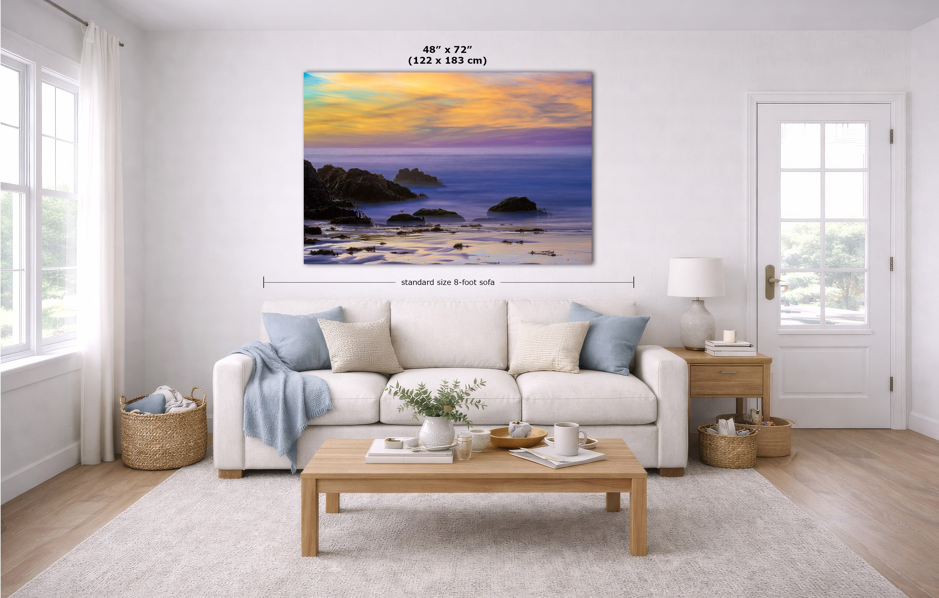 A large rectangular coastal sunset wall art print hangs above a white sofa in a bright living room. The artwork shows a beach or rocky shoreline at dusk with blue, purple, yellow, and orange tones.