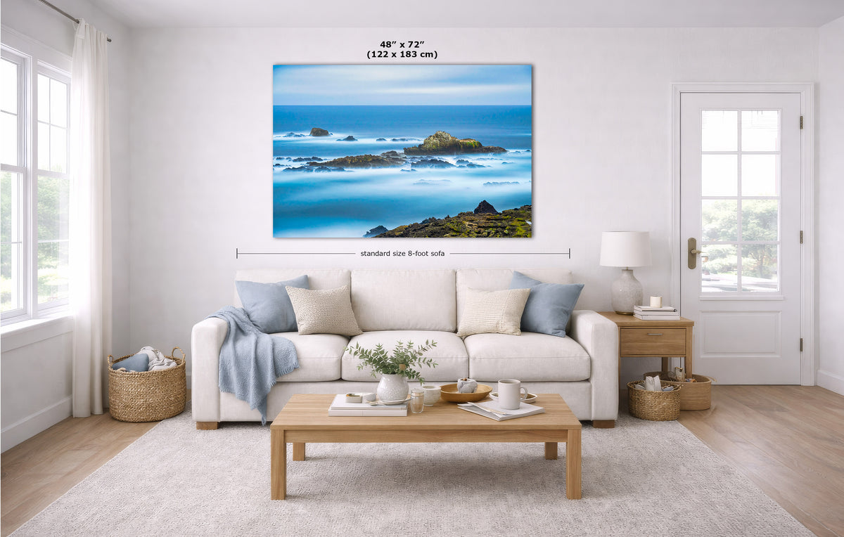 A large coastal landscape wall art print hangs above a white sofa in a bright living room. The artwork shows blue ocean waves, rocky shoreline, and bright sky.