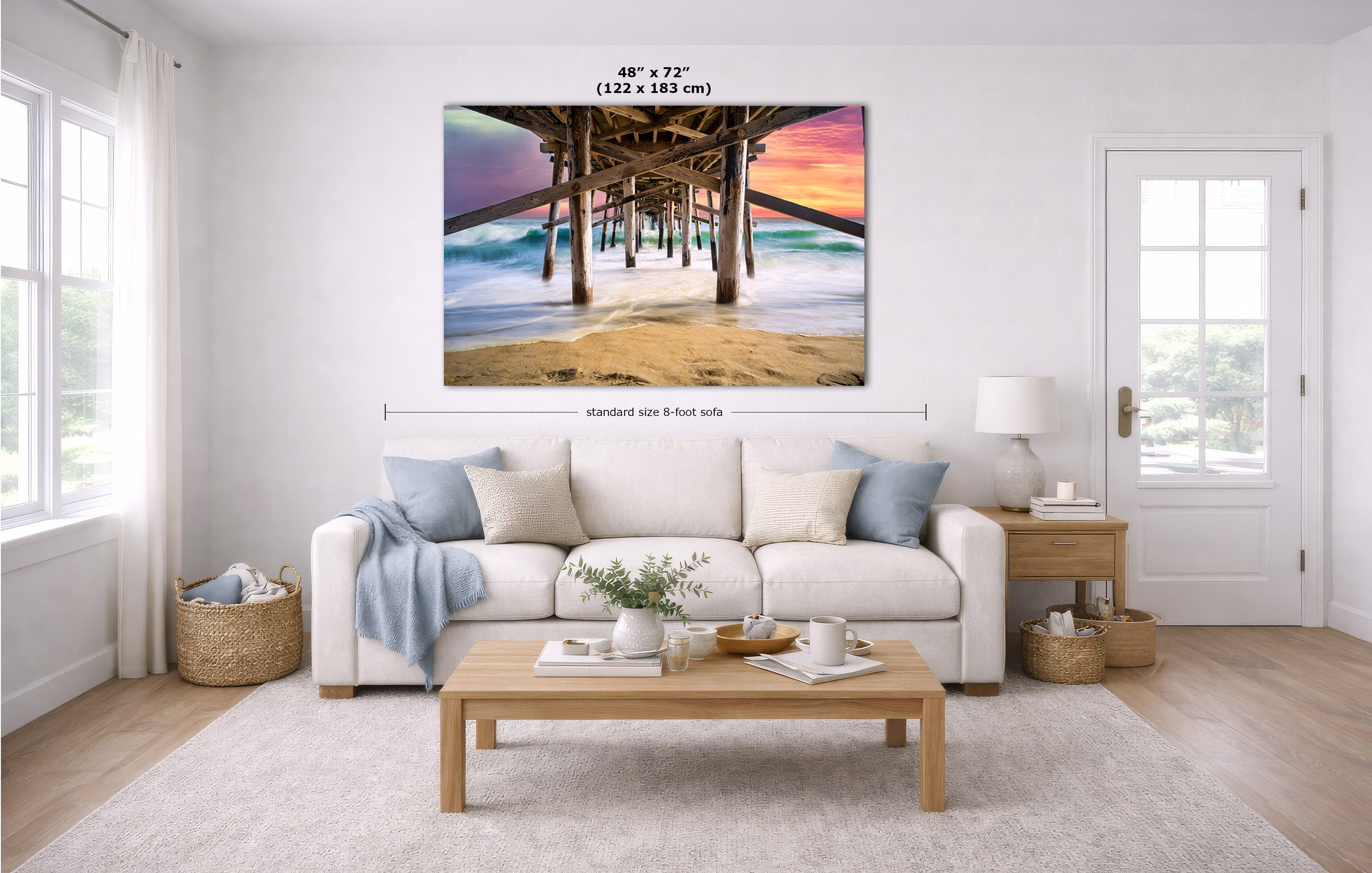 A large wall art print showing a pier over ocean waves at sunset, displayed above a white sofa in a bright modern living room. White text above the artwork indicates dimensions, and a horizontal measurement line notes standard 8-foot side spacing.