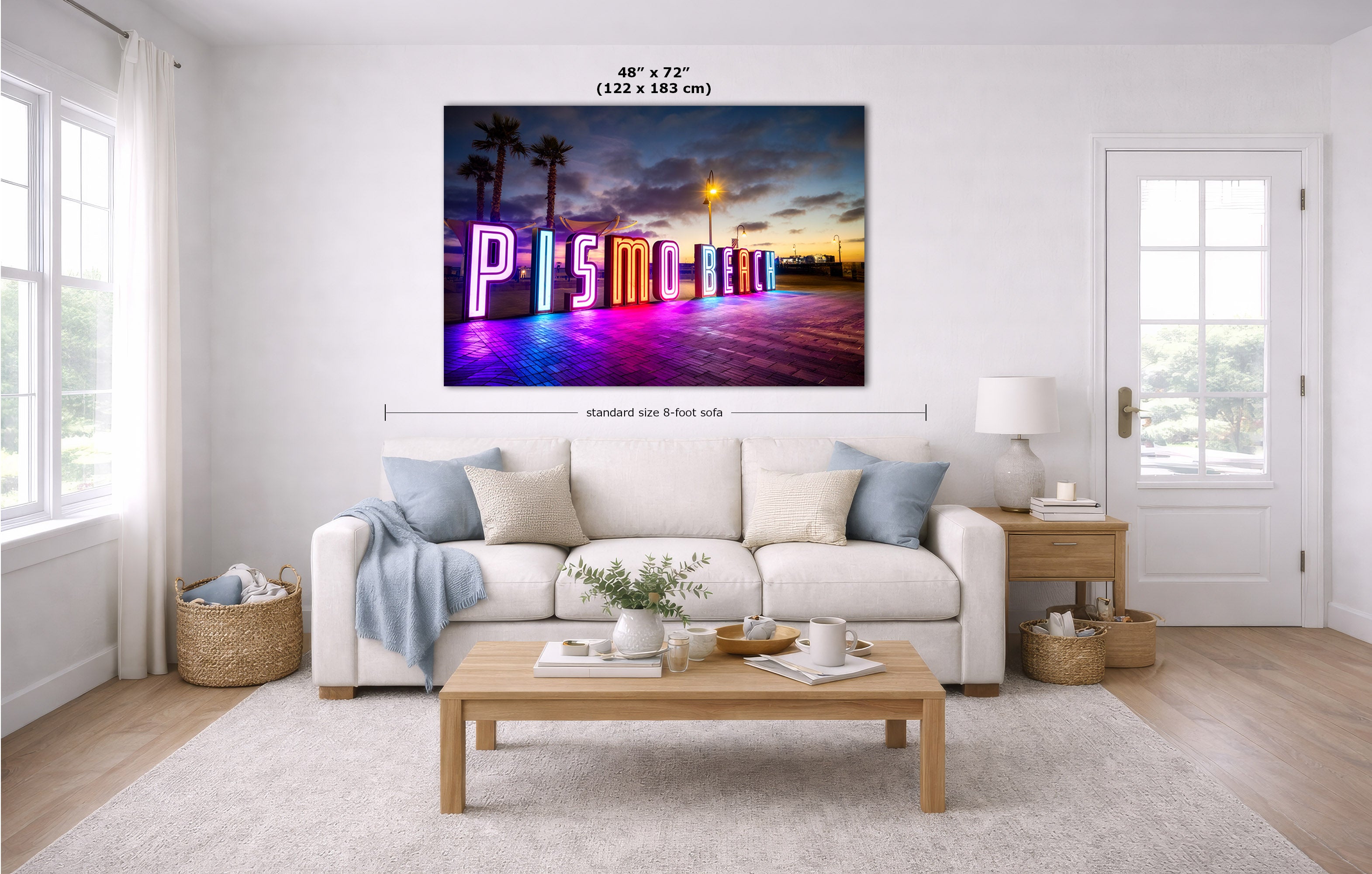 A wall art print showing a colorful neon 'Pismo Beach' sign at sunset, displayed above a white sofa in a bright modern living room. Measurement labels for the print size are overlaid on the image.