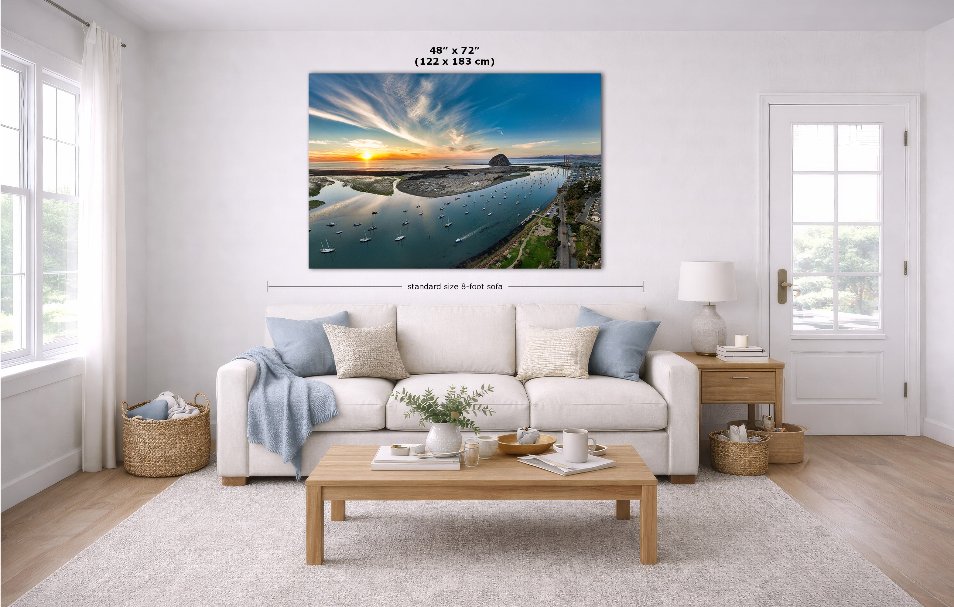 A large rectangular wall art print showing a coastal sunset over water, displayed above a white sofa in a bright modern living room. Measurement lines and text indicate the print size as 48 x 72 inches.