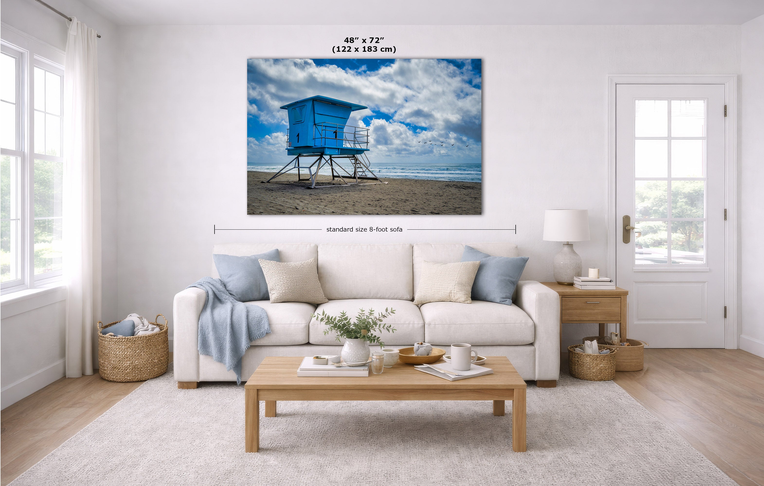 A beach scene print showing a blue lifeguard tower on a sandy shore under a cloudy sky, displayed above a white sofa in a bright modern living room. Measurement lines and text indicate the print size as 48 x 72 inches.