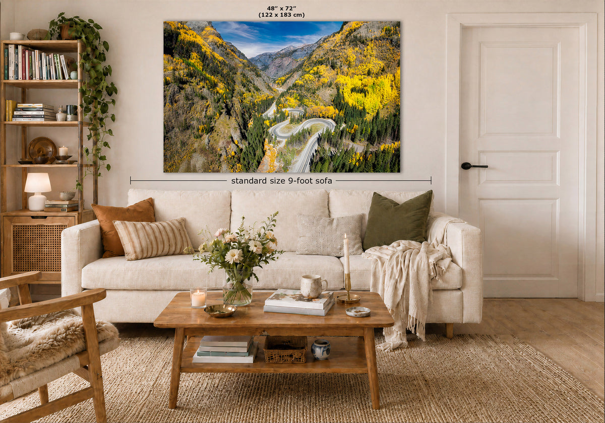 A large wall art print showing a colorful mountain valley landscape with yellow foliage and a waterfall, displayed above a white sofa in a styled living room. Measurement text indicates the print is 48 x 72 inches, and a dimension line notes standard size 9-foot sofa placement.