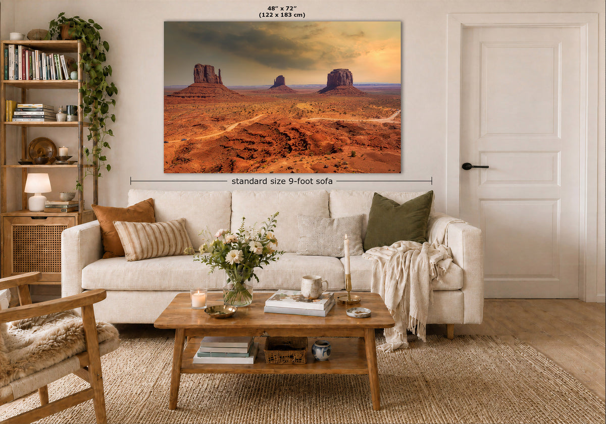 A large wall art print showing a desert landscape with red rock buttes under a warm sky, displayed above a white sofa in a styled living room. Text on the image indicates the print size as 48 x 72 inches and notes standard size 9-foot sofa placement.