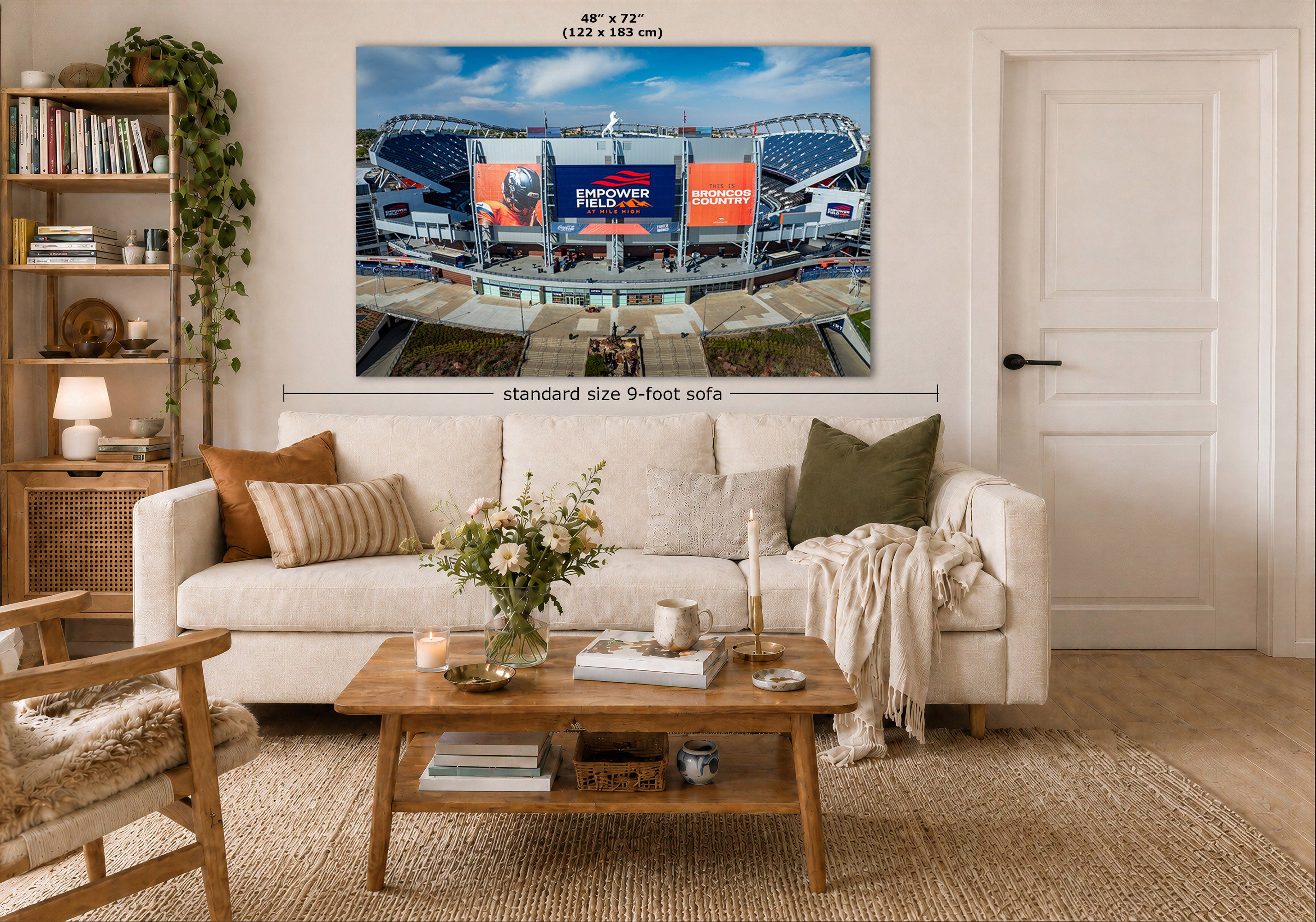 A large wall art print of Mile High football stadium in Denver, Colorado, which is displayed above a white sofa in a styled living room. The print includes a dimension label indicating 48" by 72" (122 by 183 cm), and the room scene includes a coffee table, bookshelf, and door.