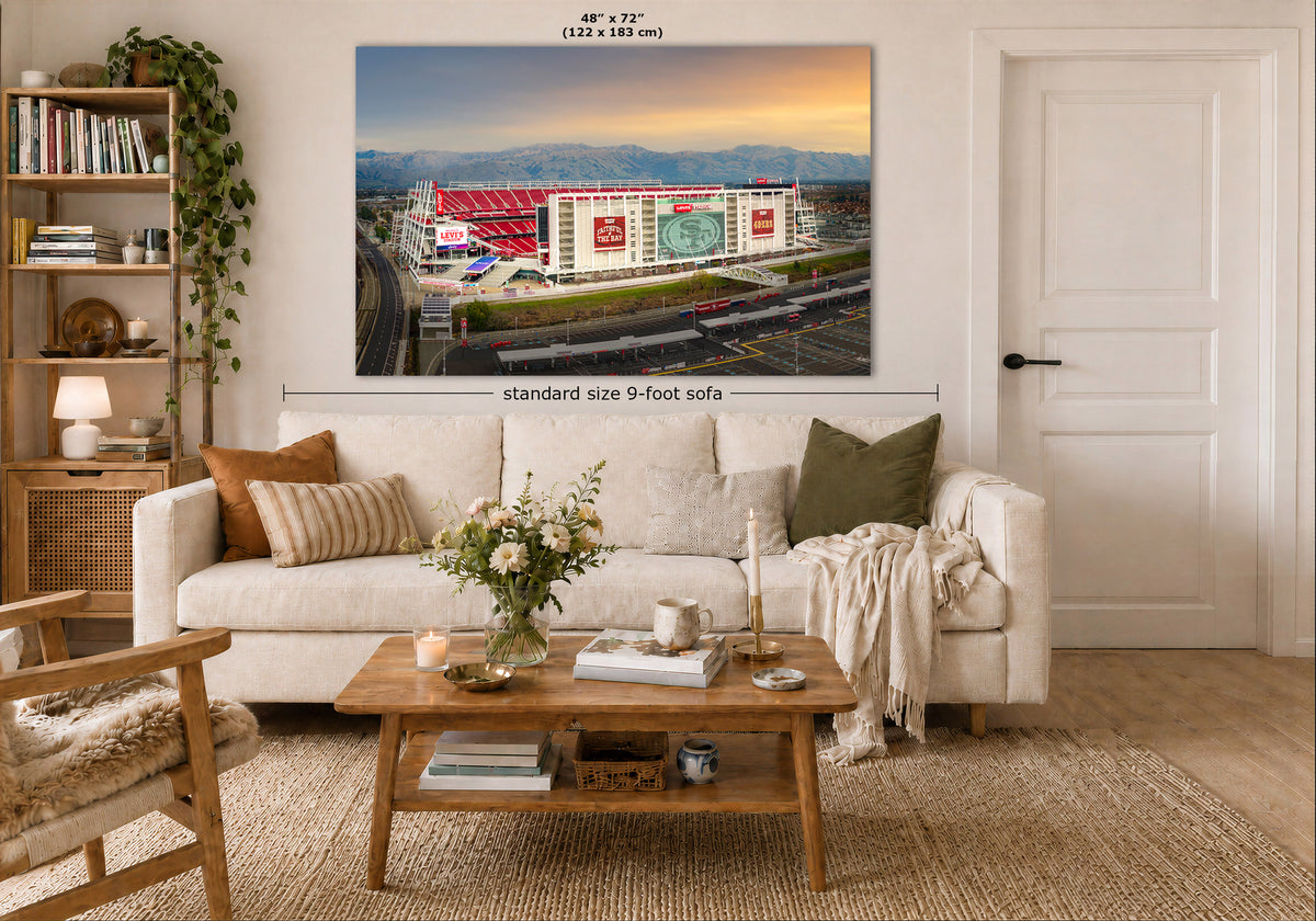 A large wall art print showing a stadium scene at sunrise, displayed above a white sofa in a styled living room. The print includes dimension labels reading 48" x 72" and a note about standard size 9-foot sofa.