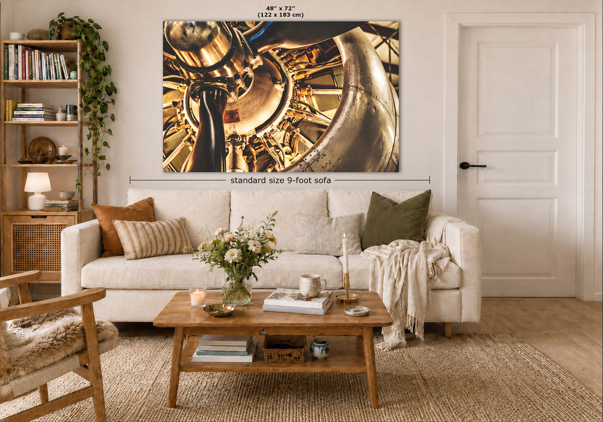 A large vintage airplane engine wall art print showing a close-up of shiny brass airplane engine parts in gold, bronze, and black tones, displayed above a white sofa in a styled living room. Measurement text and arrows indicate the print size as 48 x 72 inches, with a note reading standard size 9-foot sofa.