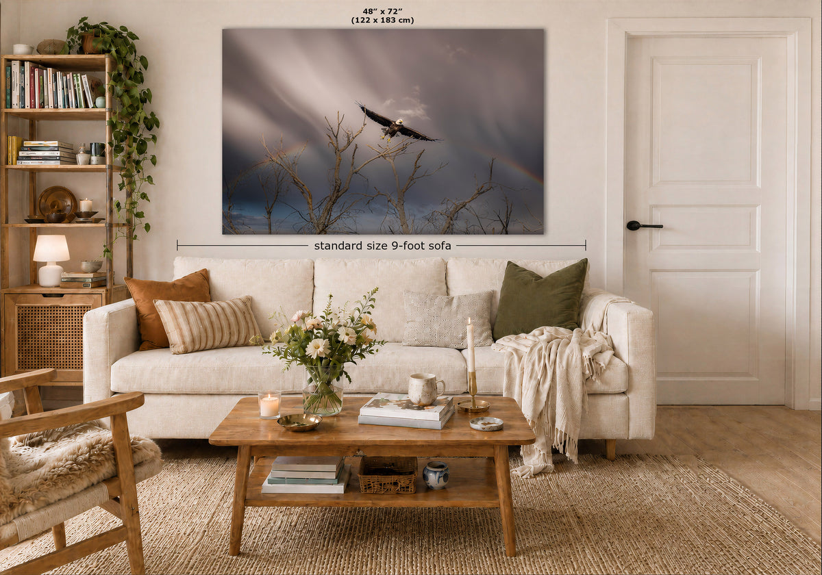 A large wall art print showing a bird flying over bare tree branches under a stormy sky with a faint rainbow, displayed above a white sofa in a styled living room. Measurement text indicates the print is 48 x 72 inches, and a dimension label notes standard size 9-foot sofa.