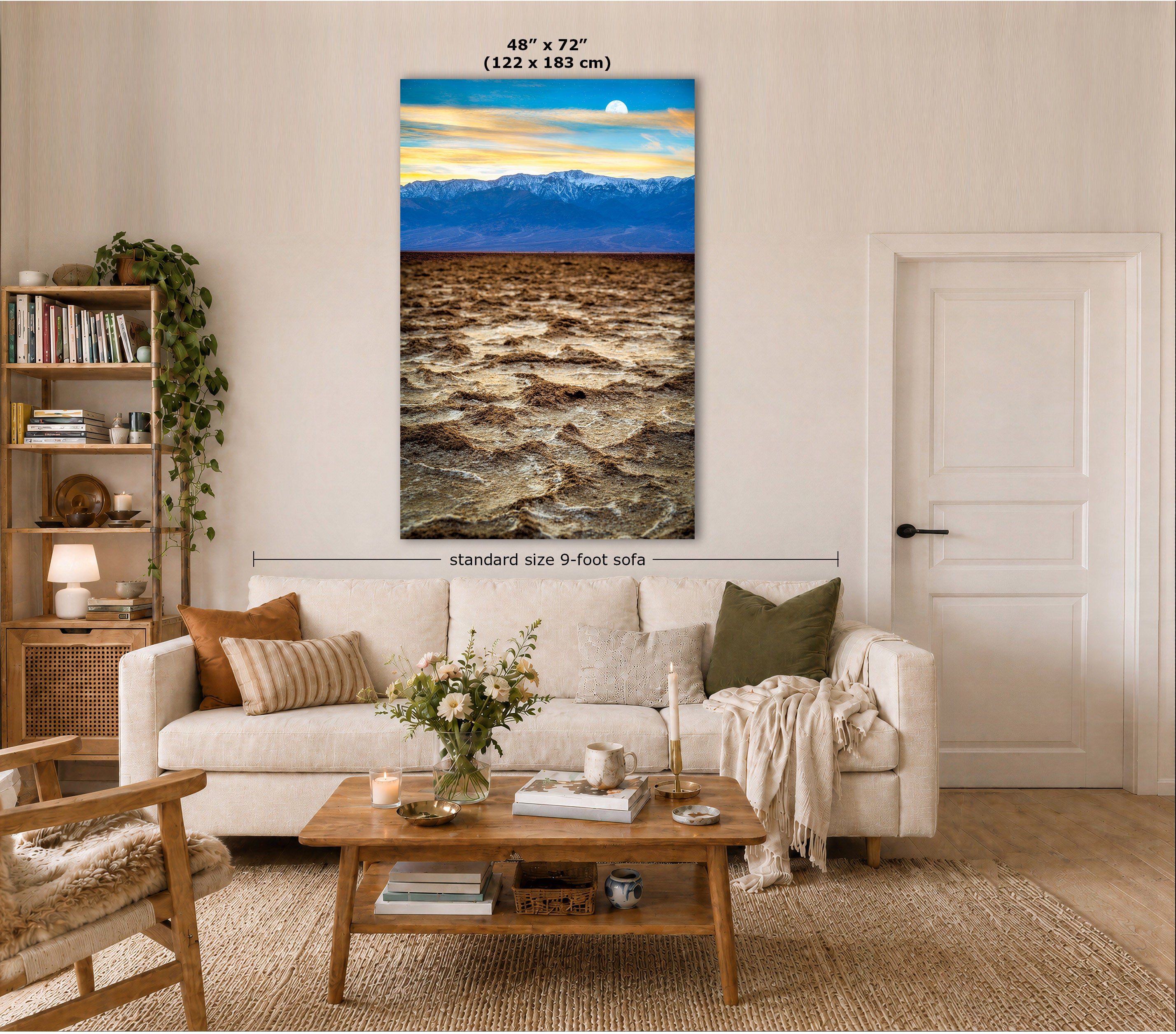 A large vertical canvas print of a dramatic mountain landscape with a glowing sunset and dark rushing river, displayed on a beige wall in a modern living room mockup above a white sofa.