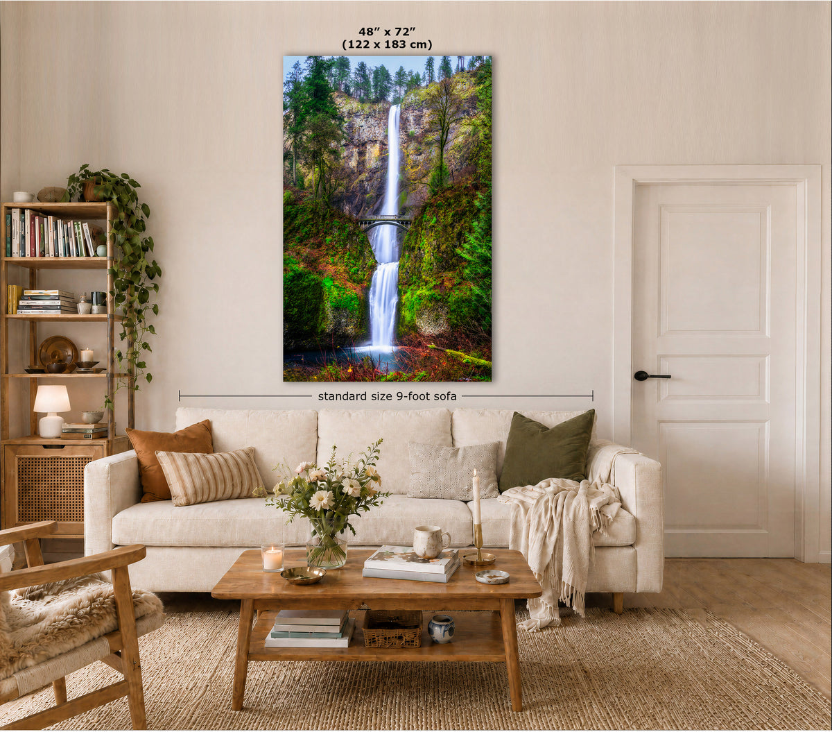 A vertical canvas wall art print of a lush green waterfall landscape is displayed on a beige wall in a modern living room setting. The print features a waterfall cascading over a rocky cliff with dense green foliage and a small bridge.