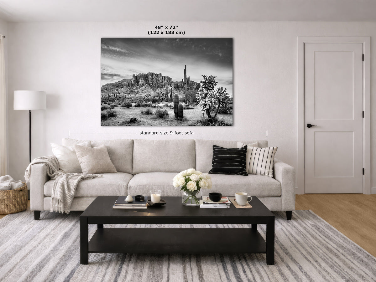 A black and white landscape canvas print of a desert scene with saguaro cacti, displayed on a wall in a modern living room mockup above a light beige sofa. The image includes dimension markers indicating the print is 48" by 72" (122 x 183 cm) wide and standard size 9-foot sofa width is shown for scale.