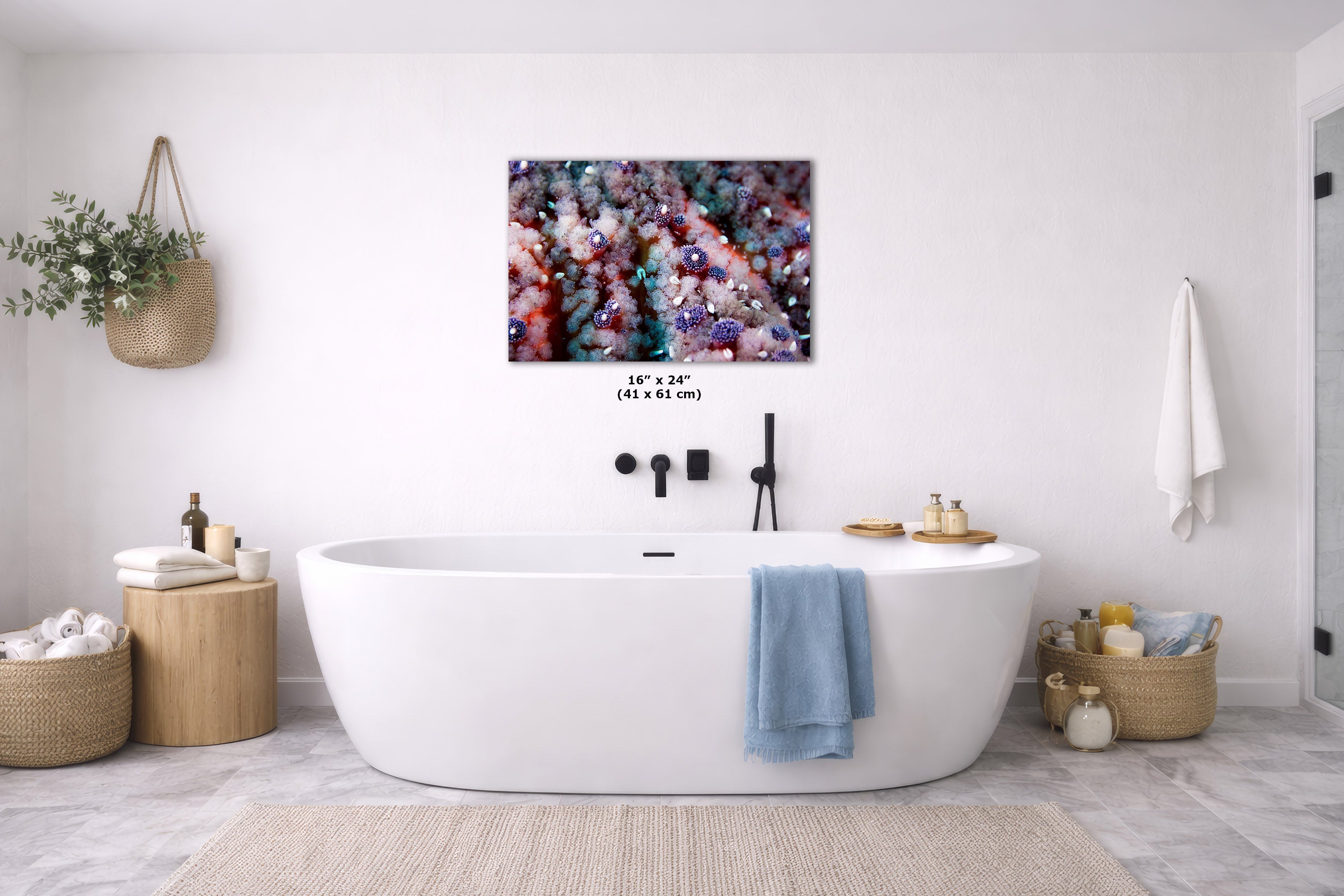 A modern freestanding bathtub in a bright white bathroom, with a colorful abstract wall art print centered above it. The scene includes a blue towel draped over the tub, woven baskets, a small plant, and a white bath mat on a light gray floor.