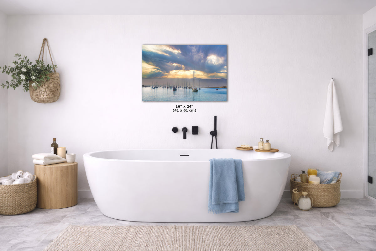 A wall art print showing a scenic sunset over calm water with boats, displayed above a white freestanding bathtub in a bright, modern bathroom. The print includes size text reading 16 x 24 in.