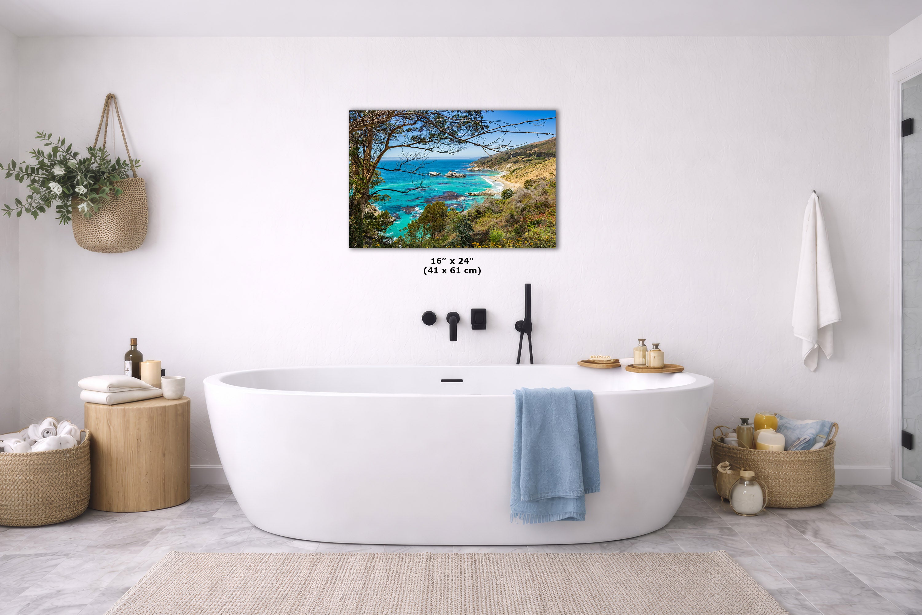 A coastal landscape wall art print hangs above a white freestanding bathtub in a bright, modern bathroom. The artwork shows turquoise water, blue sky, and green-brown shoreline.
