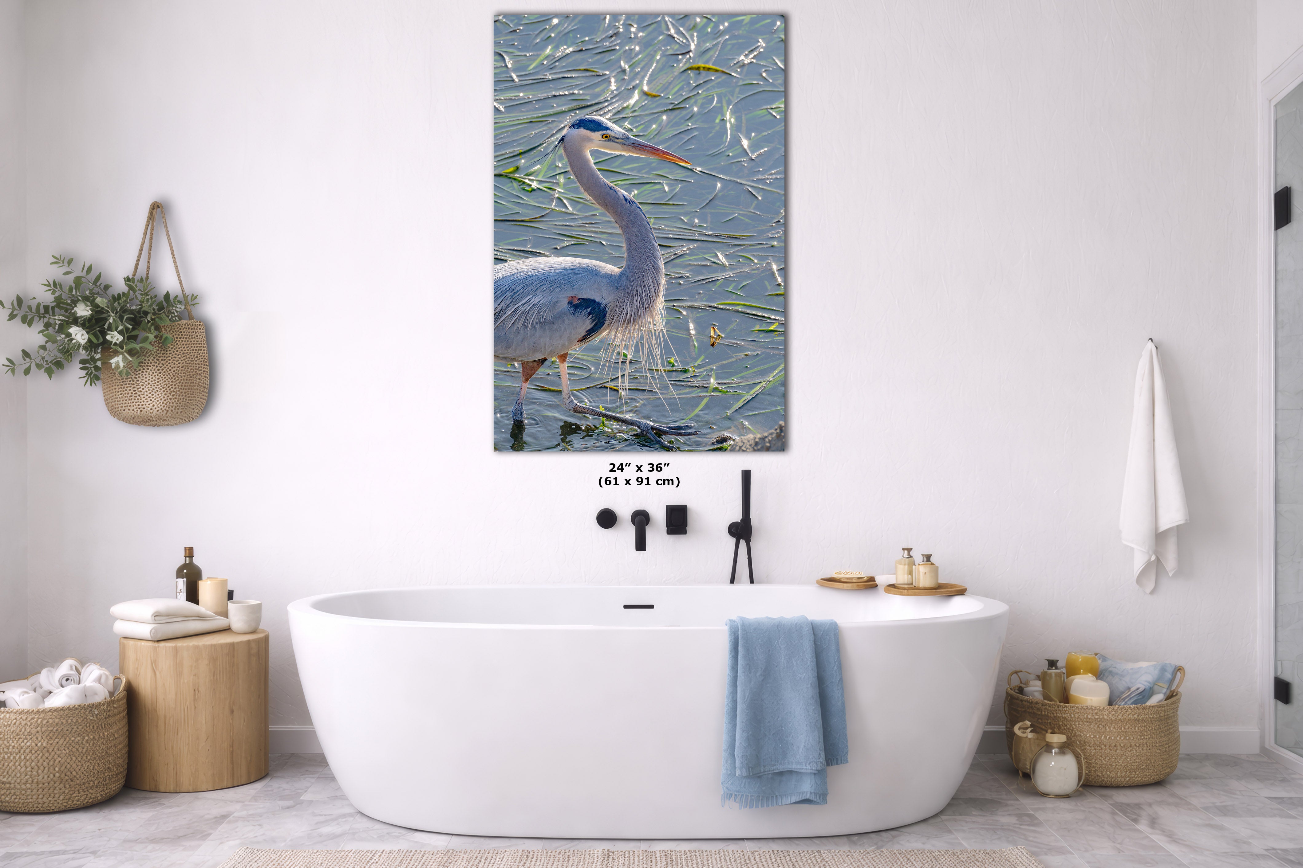 A framed wall art print depicting a blue heron standing in shallow water, displayed above a white freestanding bathtub in a bright, modern bathroom with neutral decor.