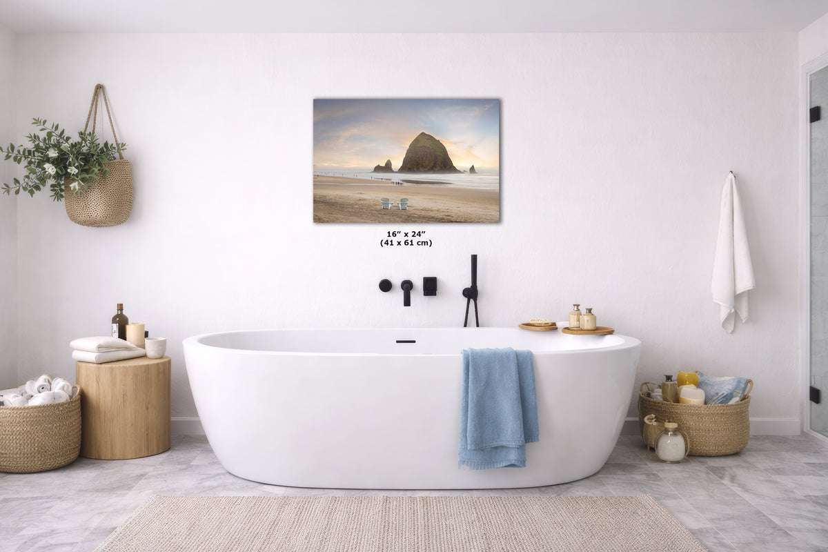 A wall art print depicting Haystack Rock at sunset is displayed above a white freestanding bathtub in a bright, modern bathroom. The print is shown with a size label of 16 x 24 inches.