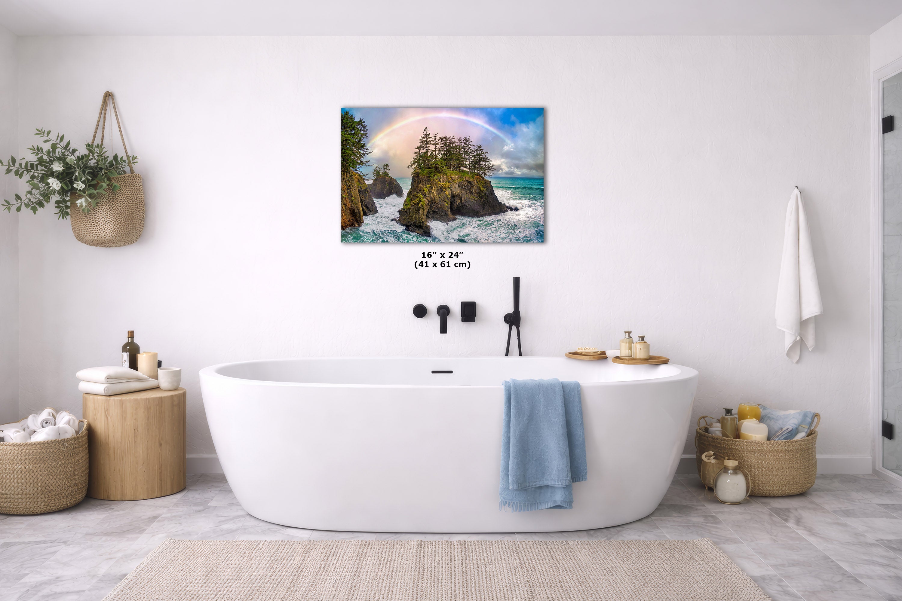 A wall art print showing a coastal landscape with rocks, ocean waves, and a rainbow, displayed above a white freestanding bathtub in a bright modern bathroom. The print includes size text reading 16 x 24 in (41 x 61 cm).