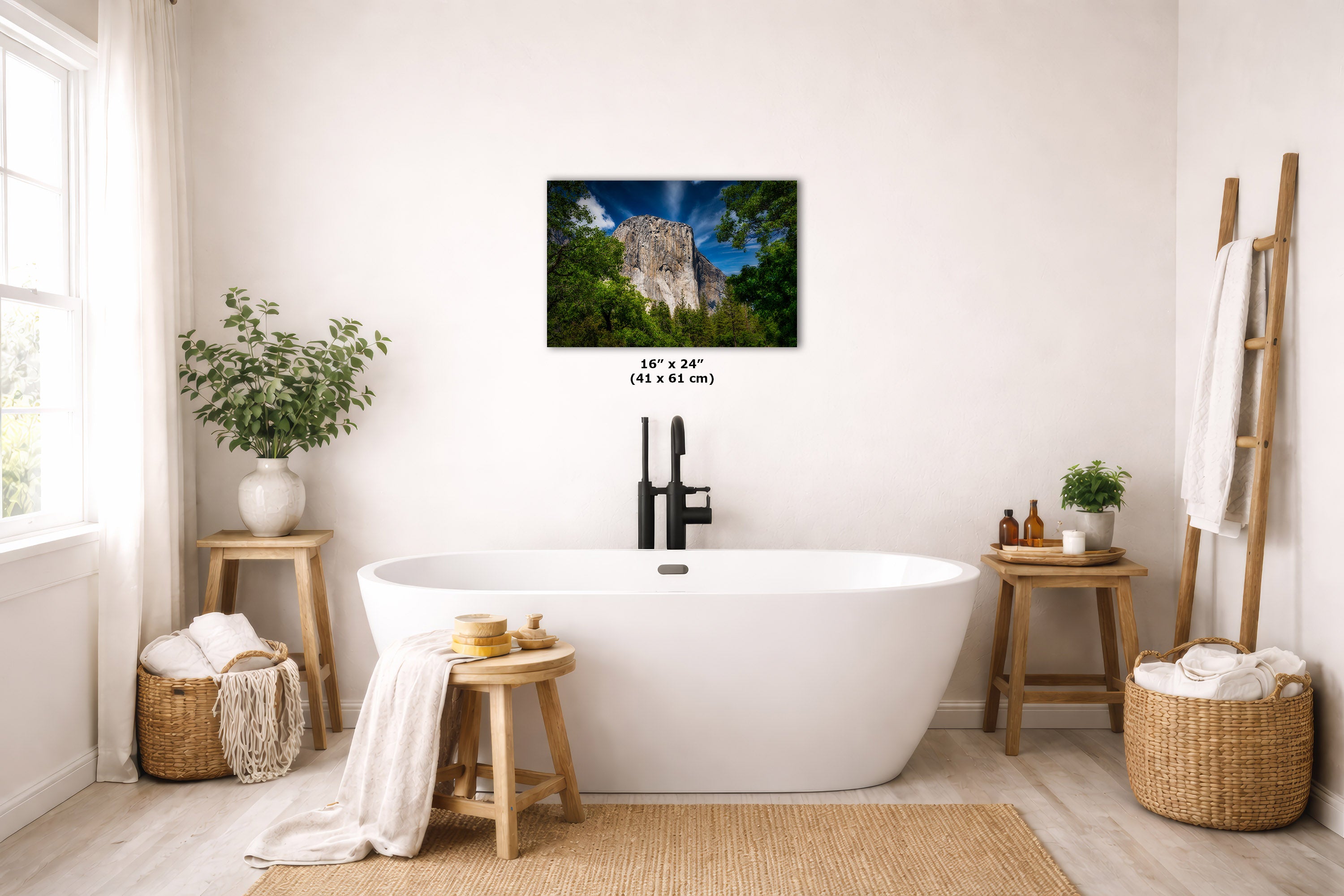 A wall art print depicting a rocky cliff and green forest hangs above a freestanding white bathtub in a bright, neutral-toned bathroom. The print includes size text reading 16 x 24 inches.