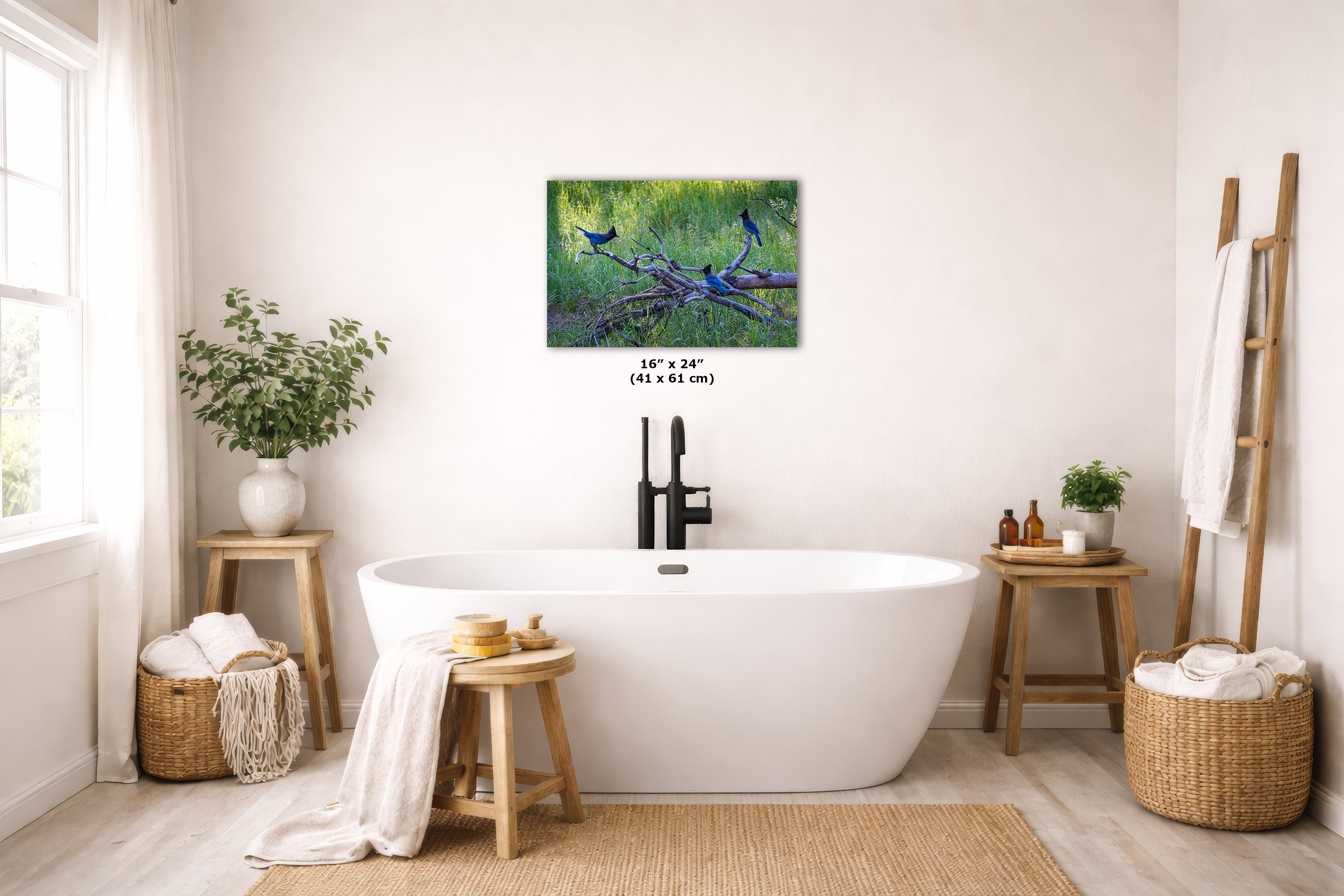 A freestanding white bathtub in a bright, neutral-toned bathroom with a framed forest wall art print above it. The scene includes wooden stools, woven baskets, plants, and a ladder-style towel rack.