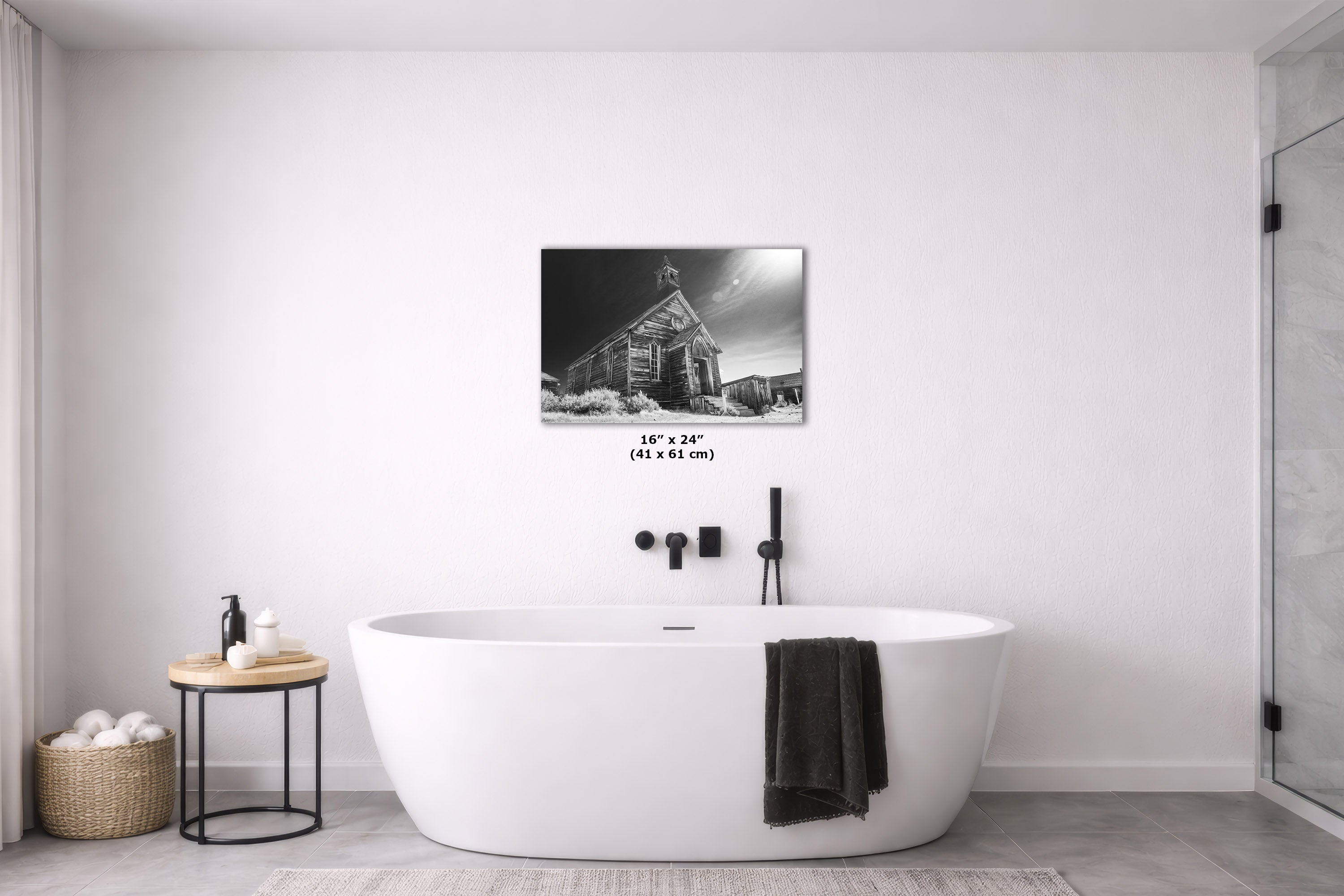 A black and white art print of a rustic cabin under a stormy sky, displayed on a white wall above a modern freestanding bathtub in a minimalist bathroom setting.