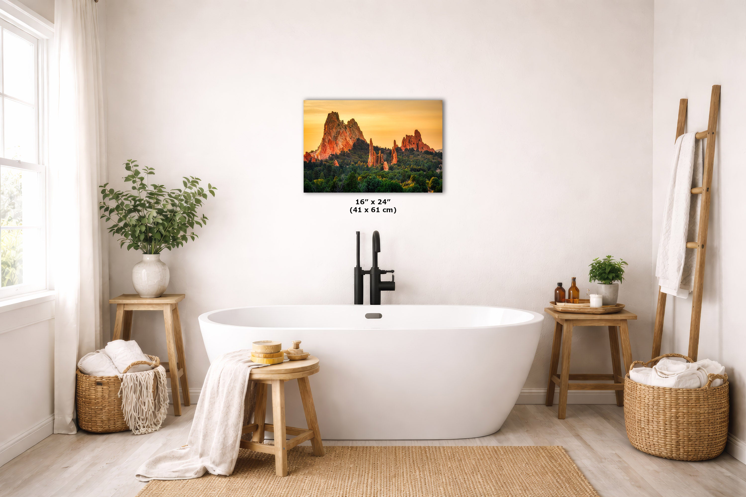 A wall art print depicting a warm sunset landscape with red rock formations is displayed above a white freestanding bathtub in a bright, modern bathroom. The print includes size text below it reading 16 x 24 inches (41 x 61 cm).