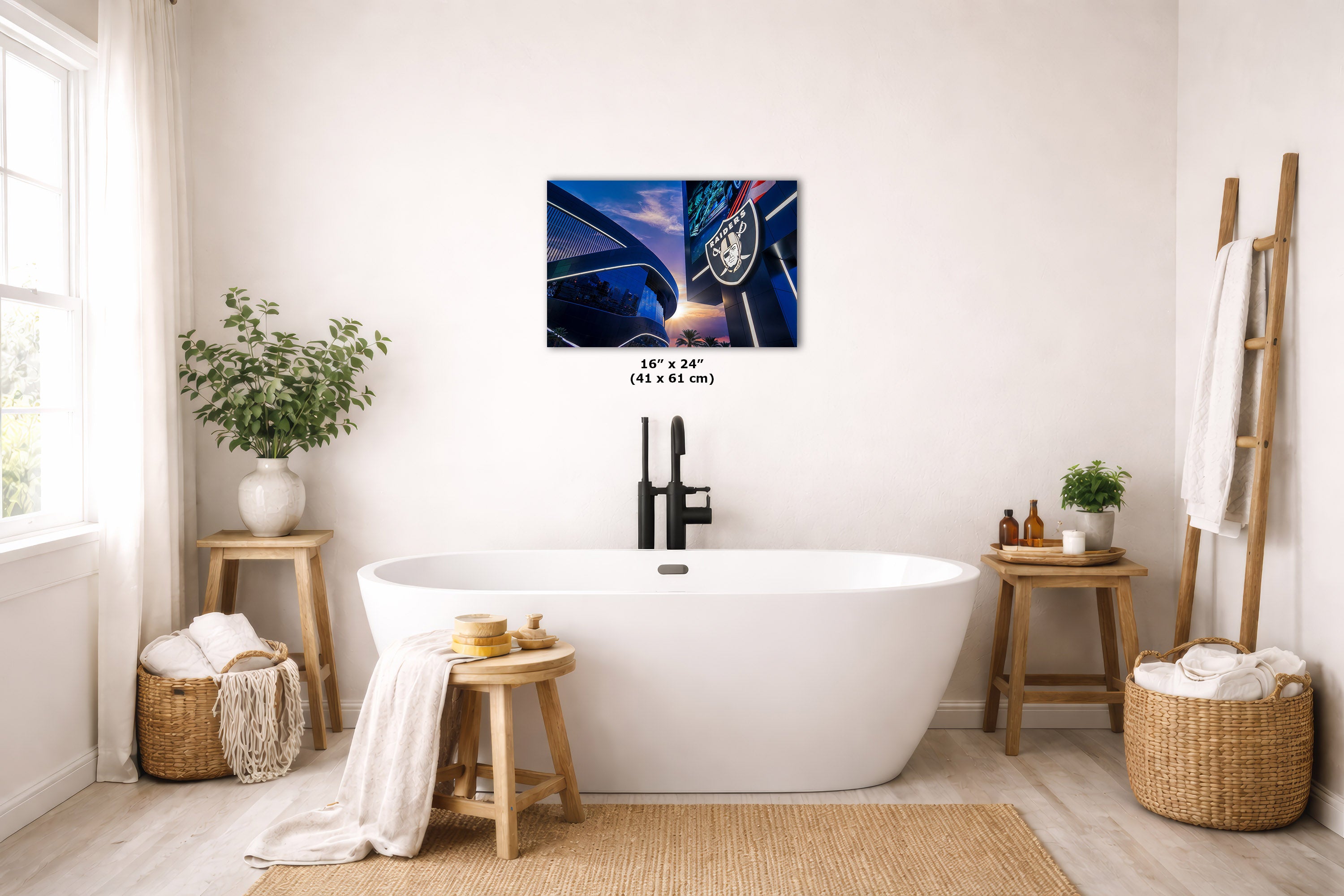 A wall art print depicting the Raider stadium hanging above a freestanding white bathtub in a bright, neutral-toned bathroom. The print includes size text reading 16 x 24 cm.