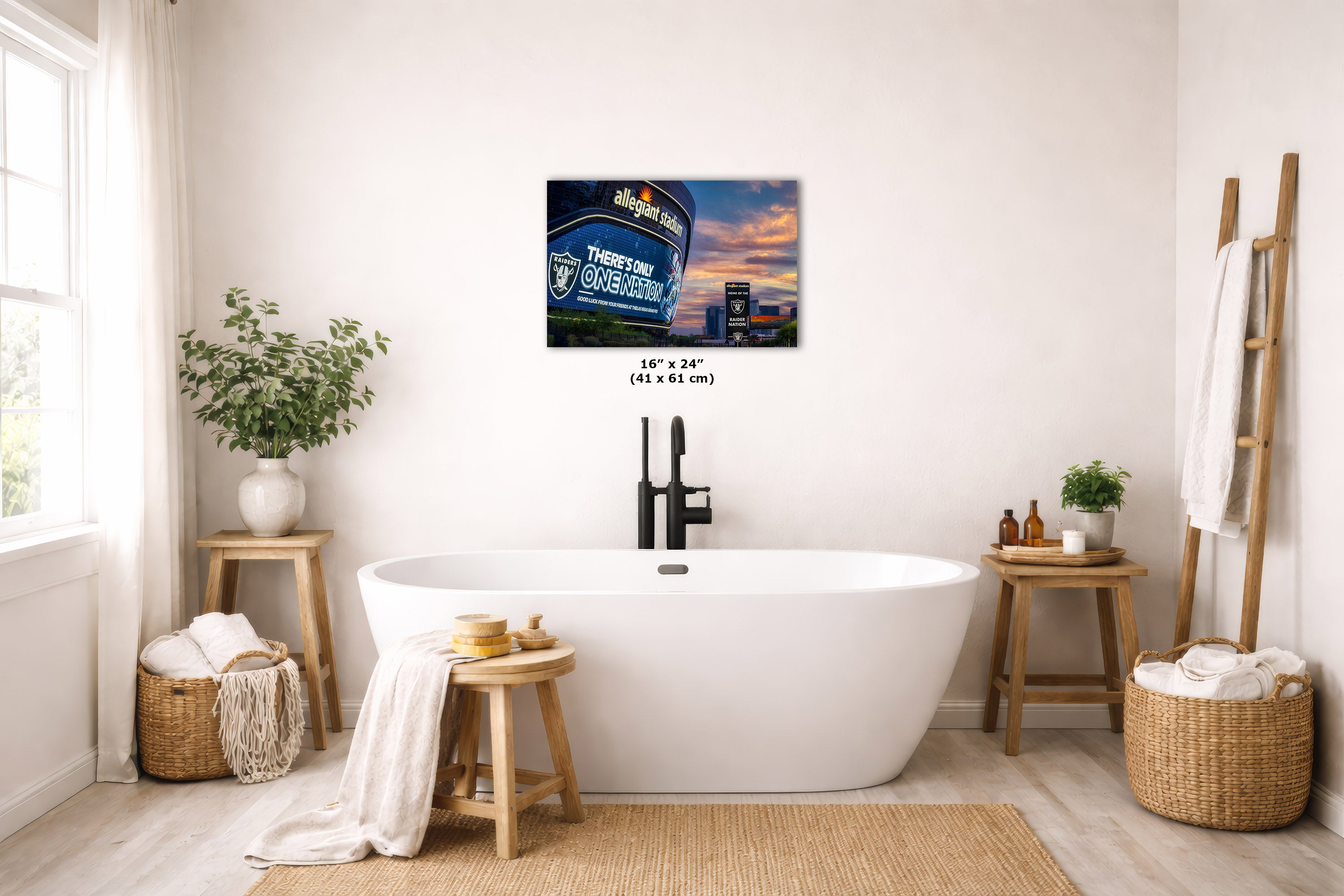 A wall art print hangs above a white freestanding bathtub in a bright, neutral-toned bathroom. The artwork depicts the Las Vegas Raiders Allegiant stadium at sunset.