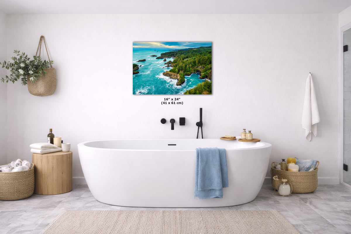 A wall art print showing a coastal landscape with turquoise water, green cliffs, and a pale sky, displayed above a white freestanding bathtub in a bright bathroom. The print includes size text reading 16 x 24 inches.