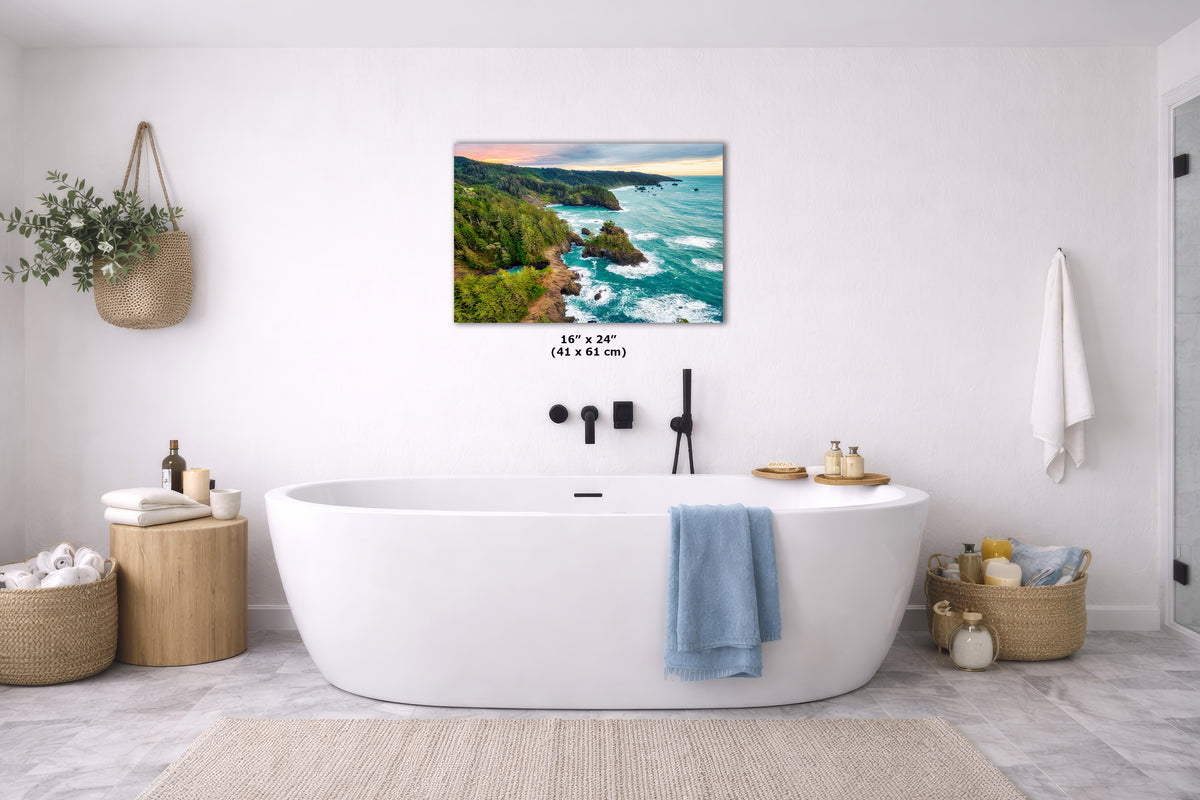 A coastal landscape wall art print hangs above a white freestanding bathtub in a bright, modern bathroom. The artwork shows a rocky shoreline with turquoise water and a sunset sky.