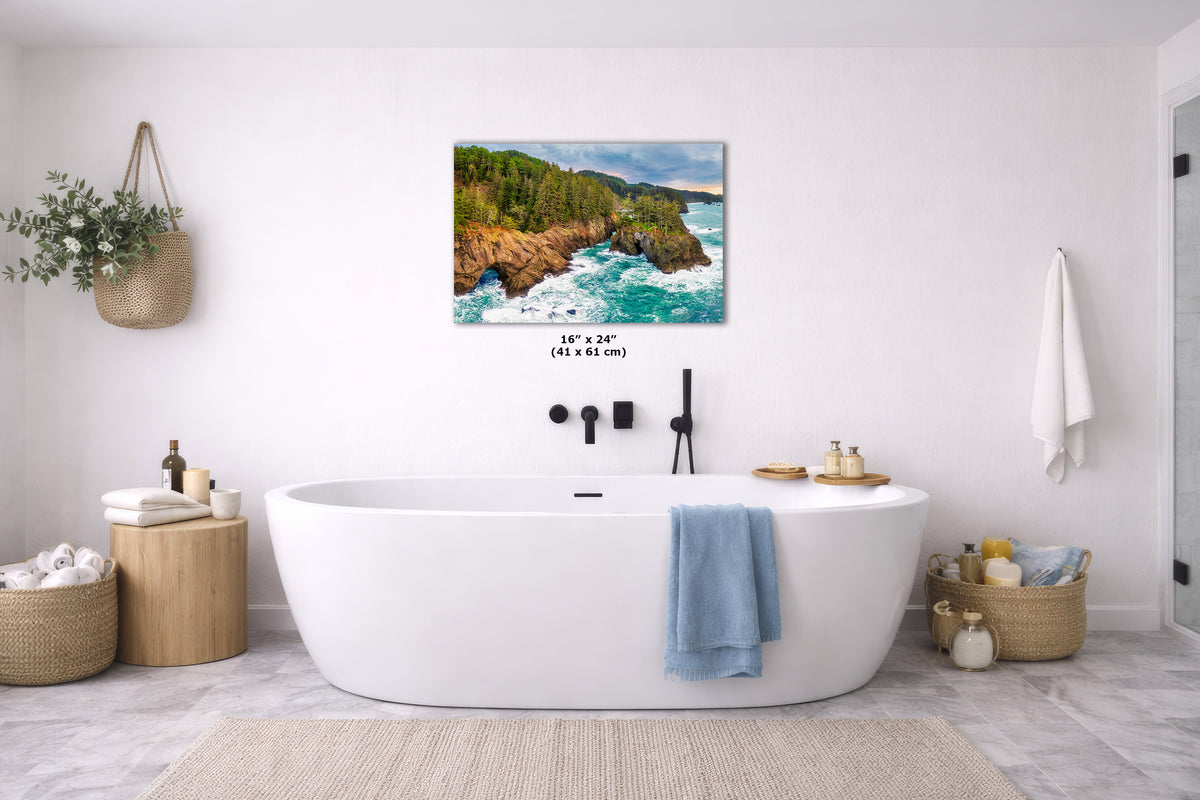 A wall art print depicting a rocky coastal shoreline with turquoise water and green trees, displayed above a white freestanding bathtub in a bright, modern bathroom. The print includes size text reading 16 x 24 in (41 x 61 cm).
