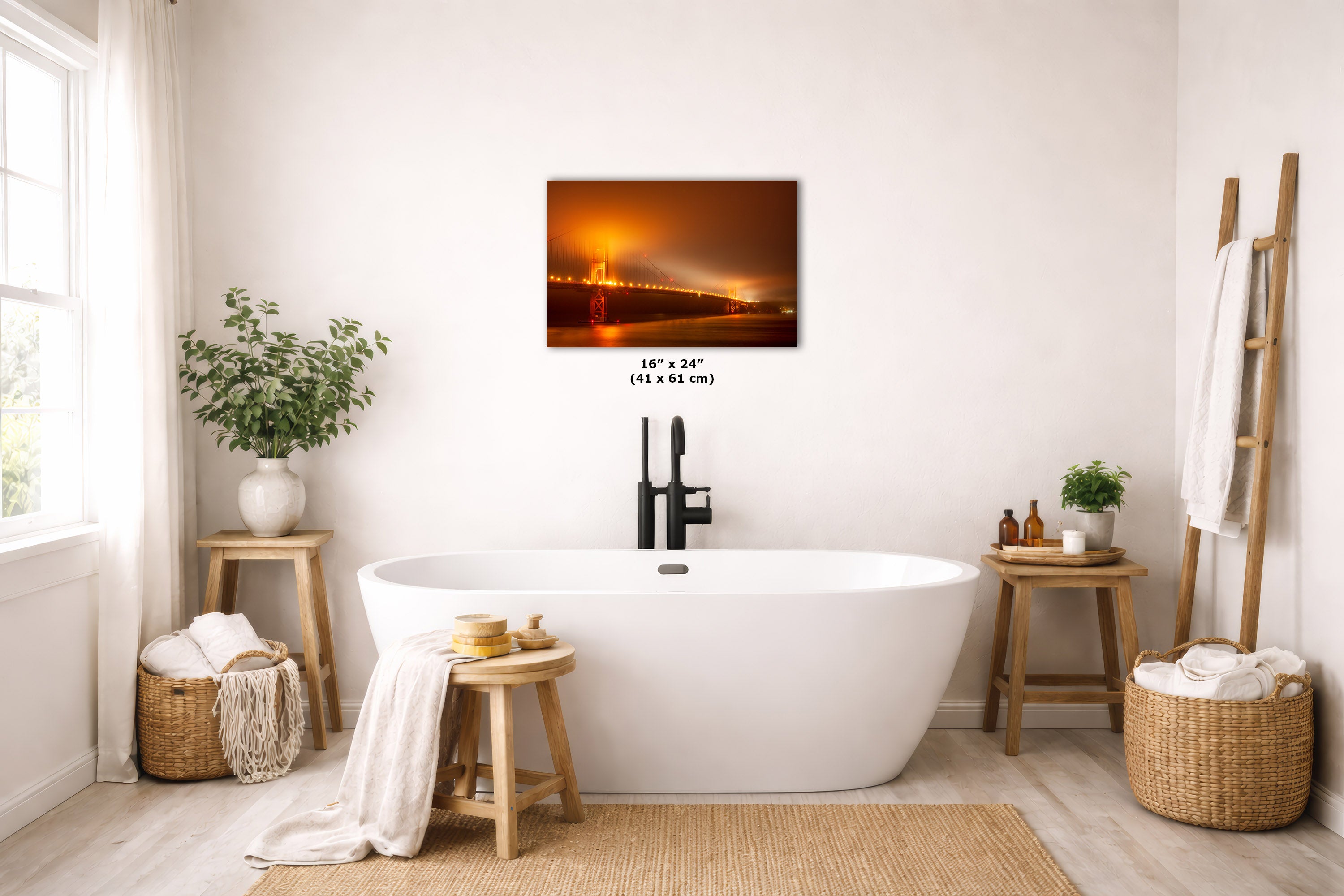 A rectangular wall art print showing a glowing orange night bridge over water, displayed above a white freestanding bathtub in a bright, neutral-toned bathroom with wood stools, baskets, plants, and a towel rack.