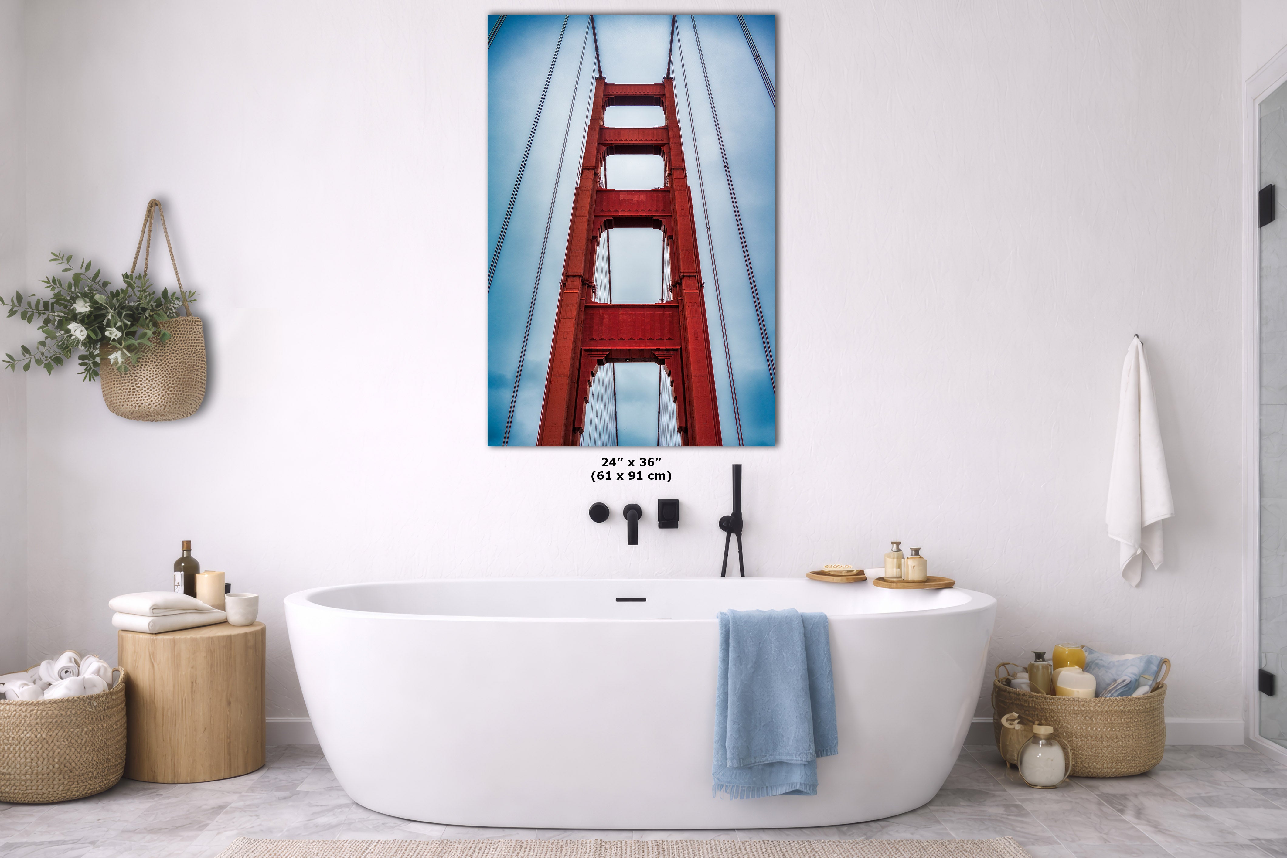 A framed wall art print of the Golden Gate Bridge hanging above a white freestanding bathtub in a bright, modern bathroom. The print shows the red bridge against a blue sky, with dimensions labeled as 24 x 36 inches.