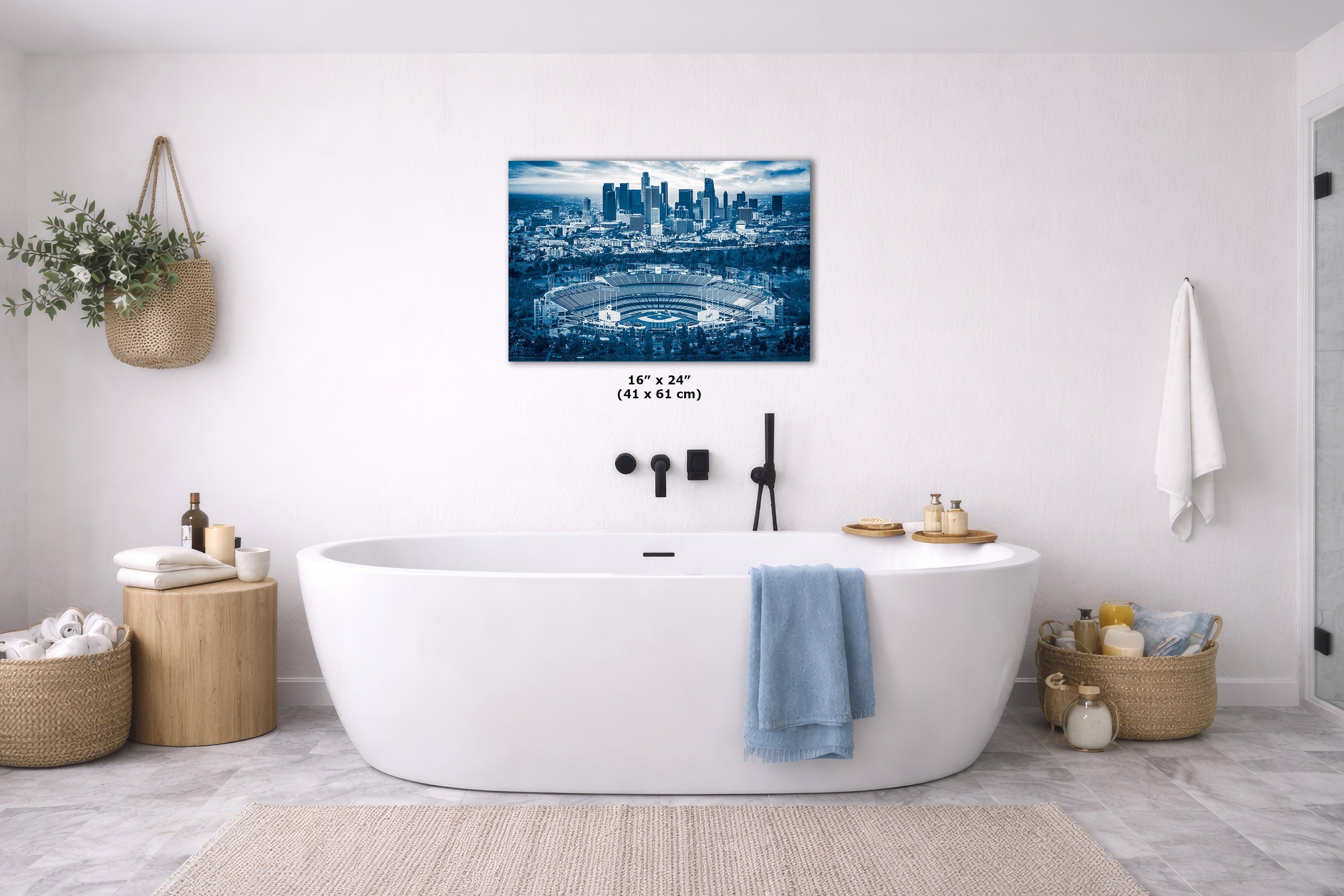 A blue-toned city skyline wall art print hangs above a freestanding white bathtub in a bright, modern bathroom with neutral decor and a blue towel draped over the tub.
