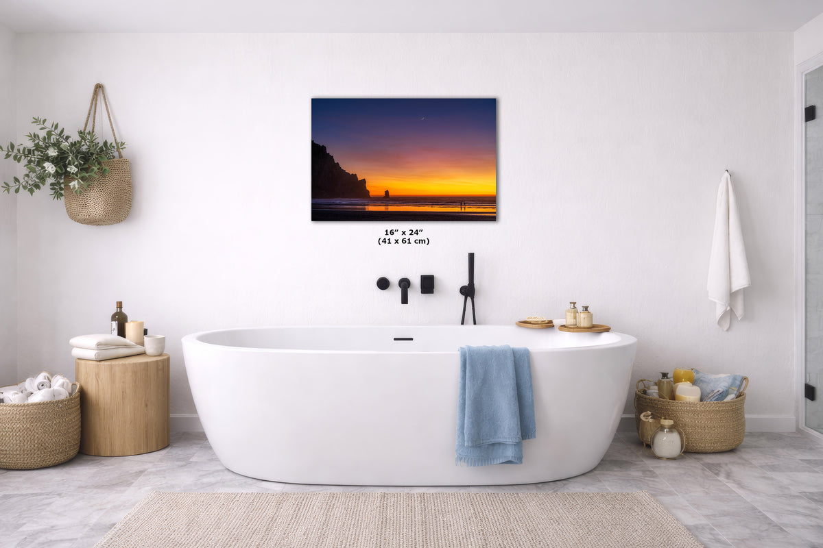 A wall art print showing a sunset over a beach cliff, displayed above a white freestanding bathtub in a bright modern bathroom. The print includes size text reading 16" x 24" (41 x 61 cm).