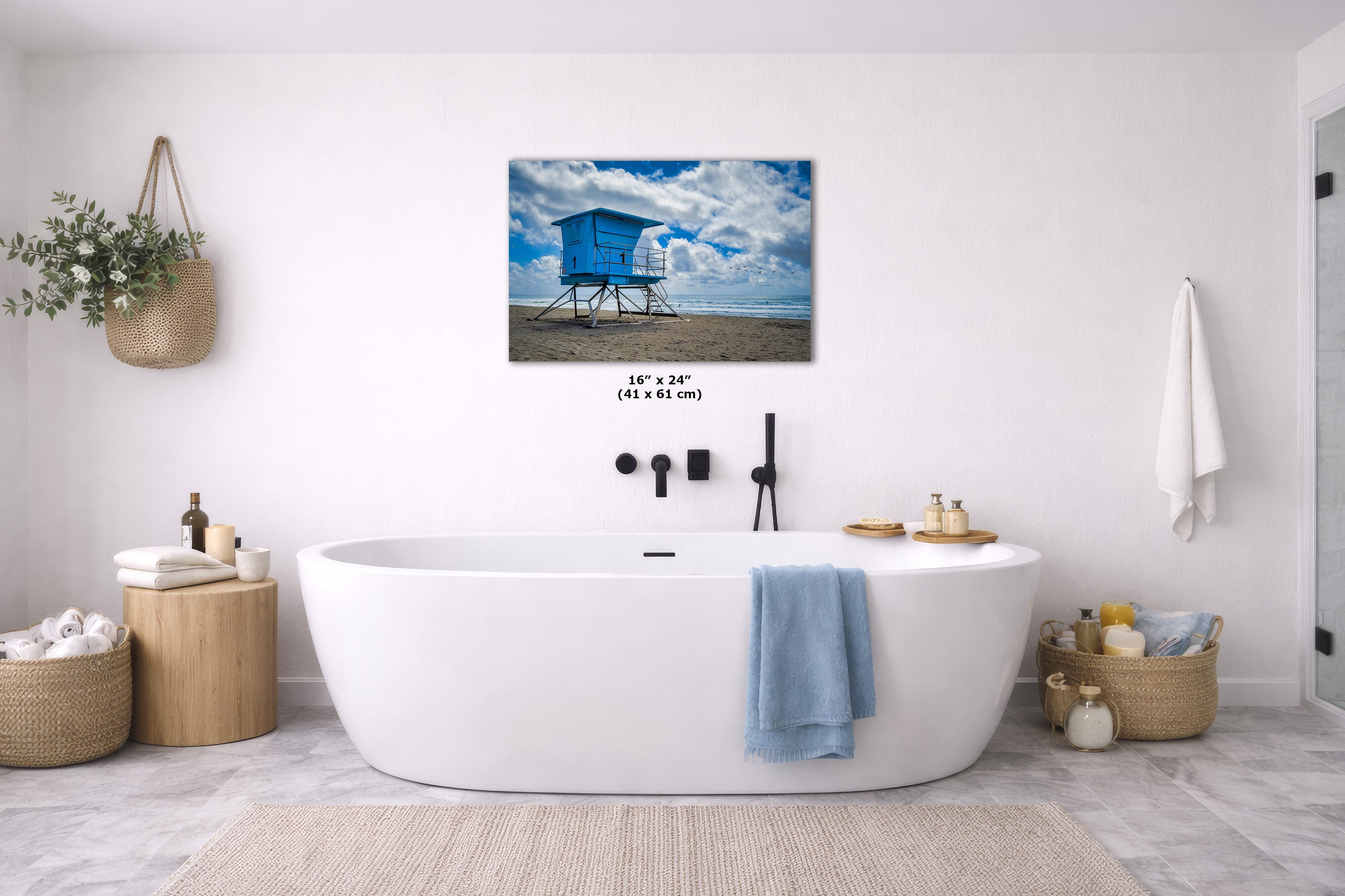 A wall art print showing a blue beach lifeguard tower under a cloudy sky, displayed above a white freestanding bathtub in a bright modern bathroom. The print includes size text reading 16 x 24 inches.