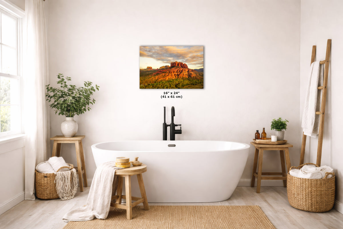 A wall art print depicting a warm sunset over red rock hills is displayed above a white freestanding bathtub in a bright, neutral-toned bathroom. The print includes size text reading 16 x 24 inches (41 x 61 cm).