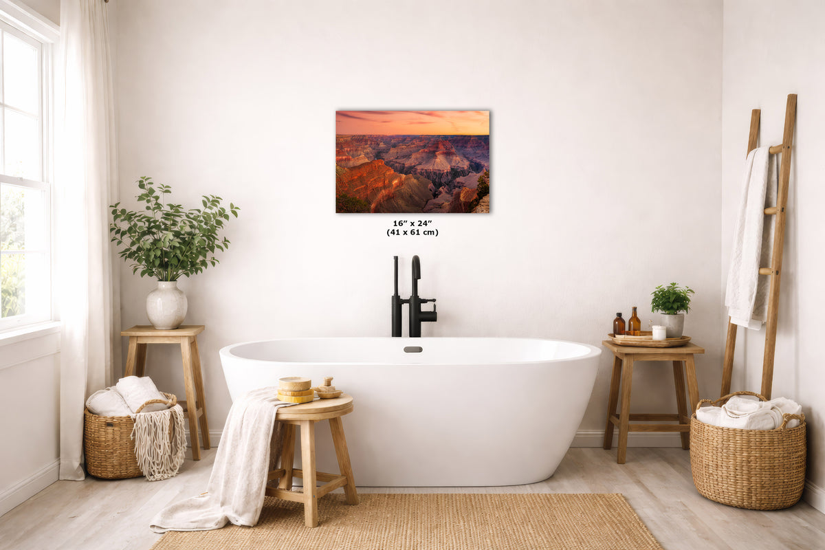 A wall art print depicting a mountain landscape with a pink and orange sunset sky, displayed above a white freestanding bathtub in a bright, modern bathroom with wooden stools, plants, and woven baskets.