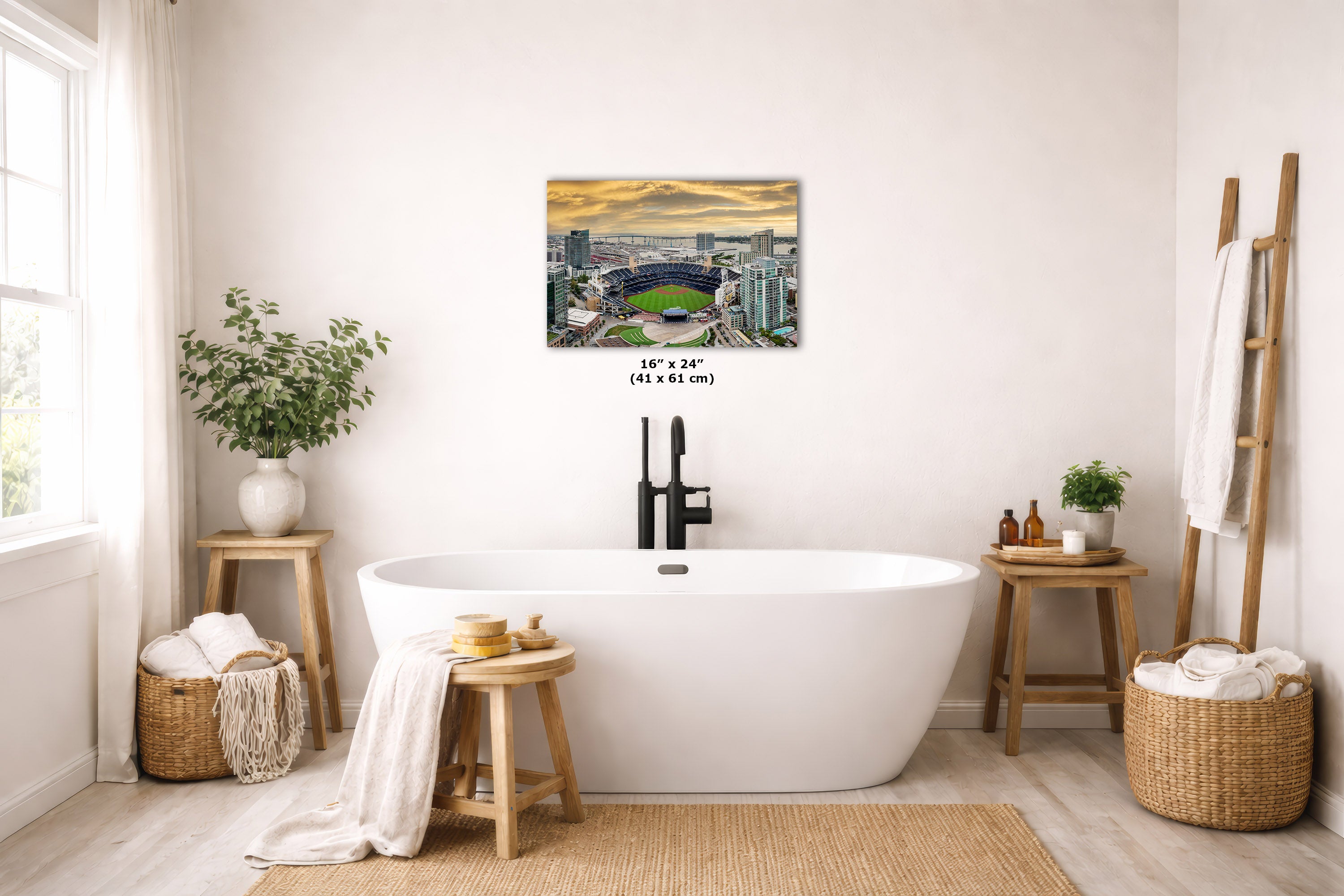 A wall art print showing a stadium at sunset hangs above a freestanding white bathtub in a bright, neutral bathroom. The print includes a small size label reading 16 x 24 inches (41 x 61 cm).