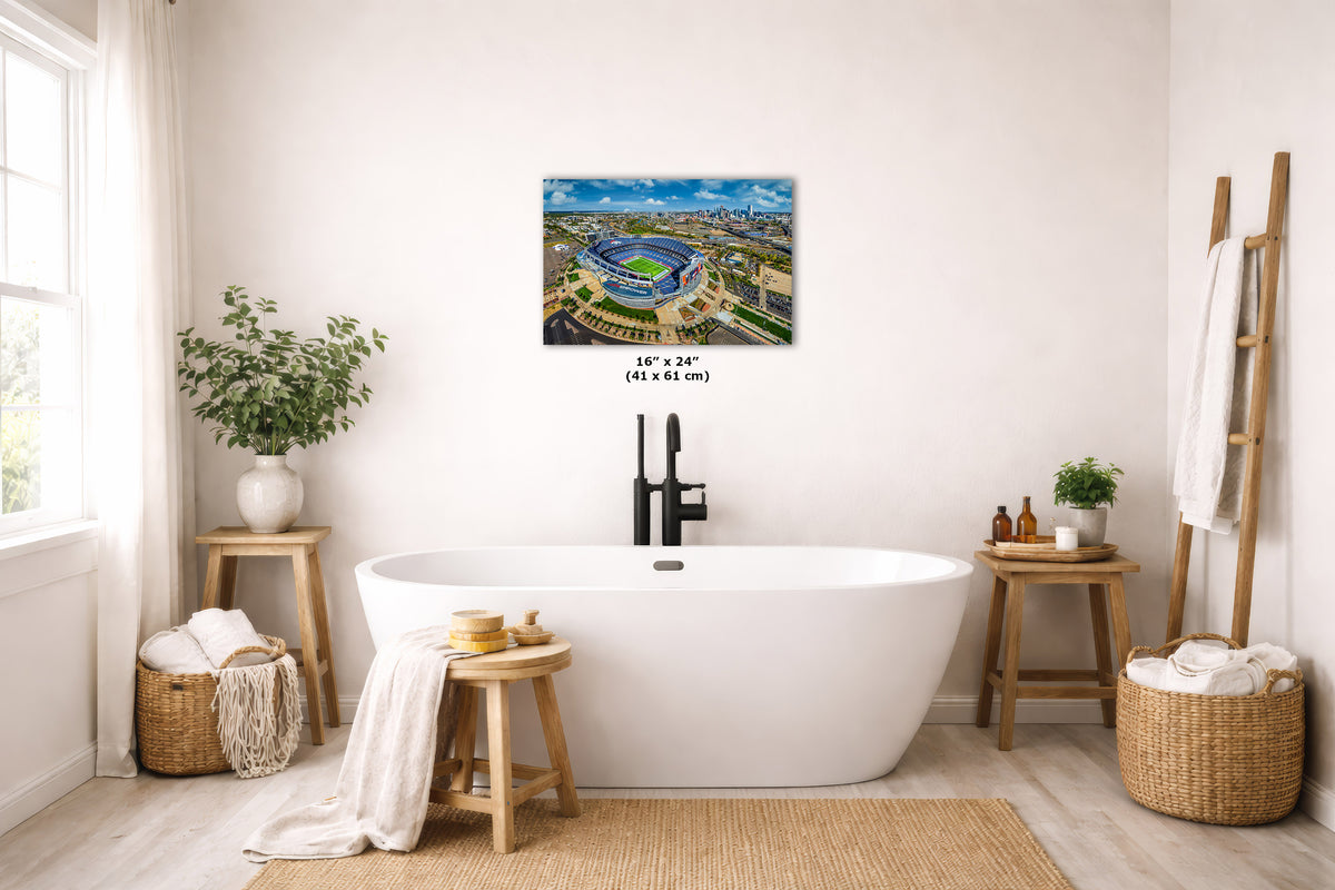 A landscape art print depicting Broncos' Mile High Stadium in Denver Colorado, displayed on a light beige wall above a white freestanding bathtub in a styled, bright bathroom setting.