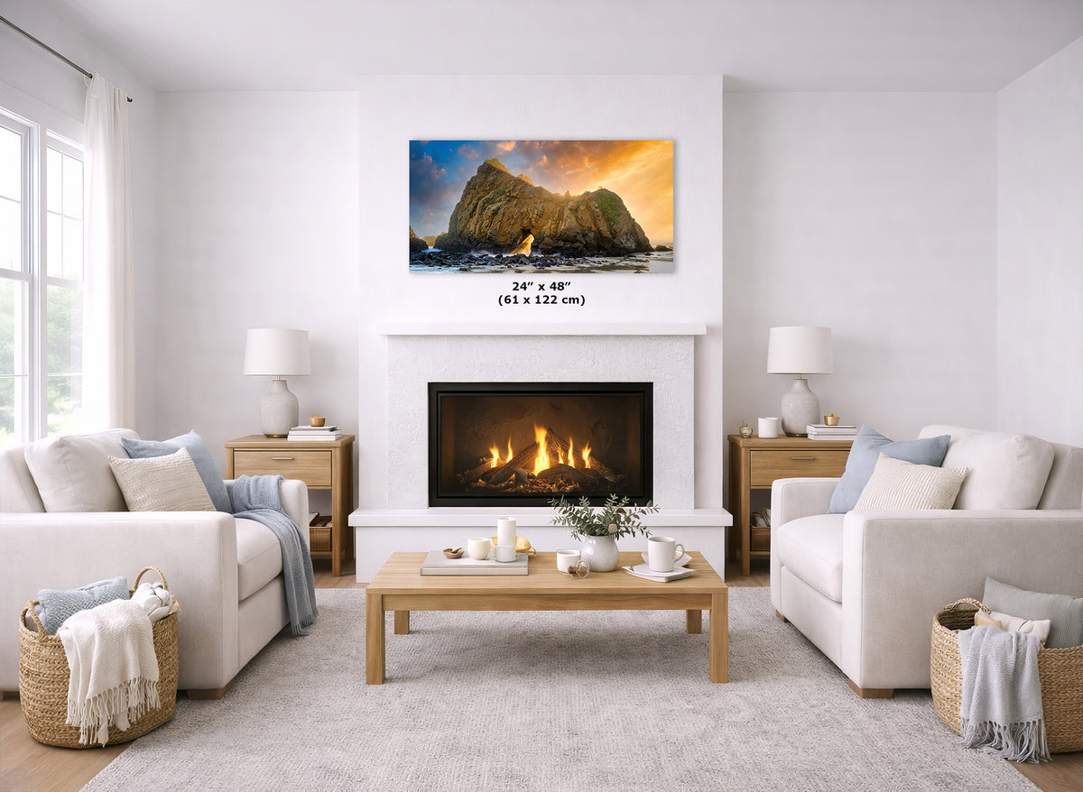 A framed wall art print depicting a dramatic rocky coastline at sunset hangs above a fireplace in a bright, modern living room. The print includes size text reading 24 x 48" (61 x 122 cm).