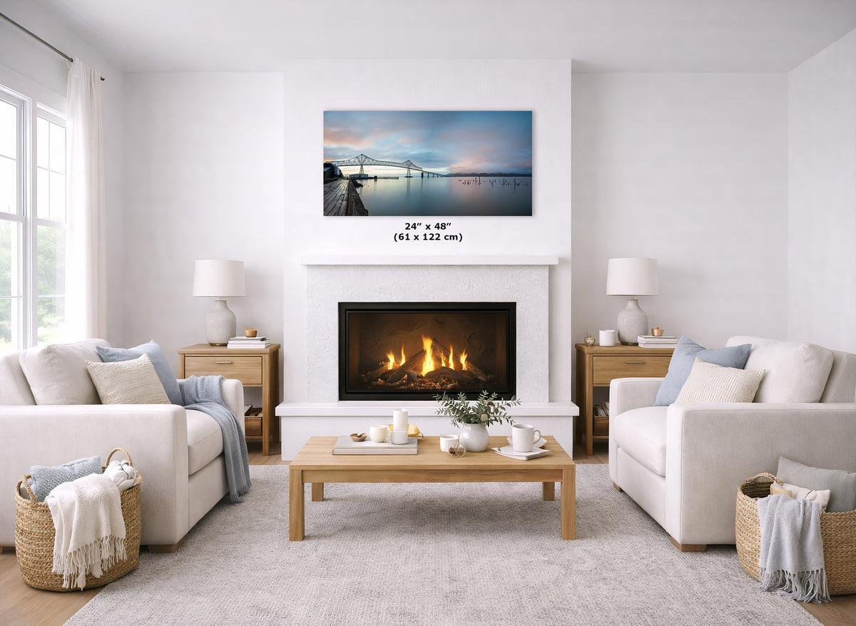 A framed landscape art print of a pier and bridge over water is displayed above a fireplace in a bright, modern living room. The print is shown with a size measurement overlay reading 24" x 48" (61 x 122 cm).