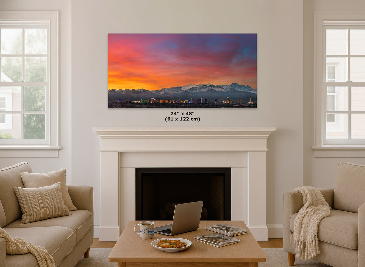 A framed wall art print showing a colorful sunset over a mountain range is displayed above a white fireplace in a bright living room. The print includes size text reading 24 x 48 inches (61 x 122 cm).