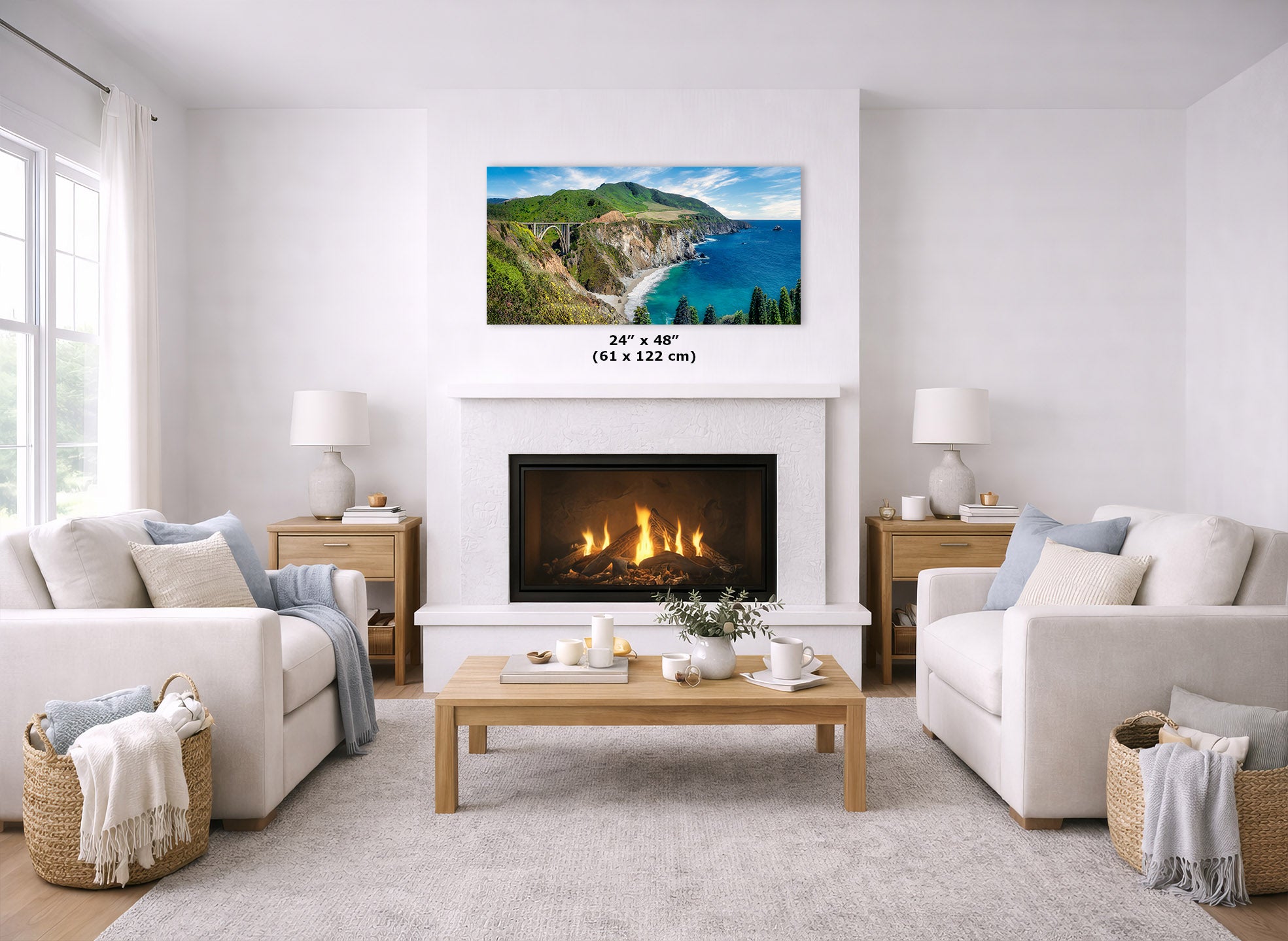 A landscape wall art print showing green cliffs and blue ocean water, displayed above a fireplace in a bright, modern living room with white sofas, a wooden coffee table, and a light rug. The print includes a size label reading 24 x 48 (61 x 122 cm).