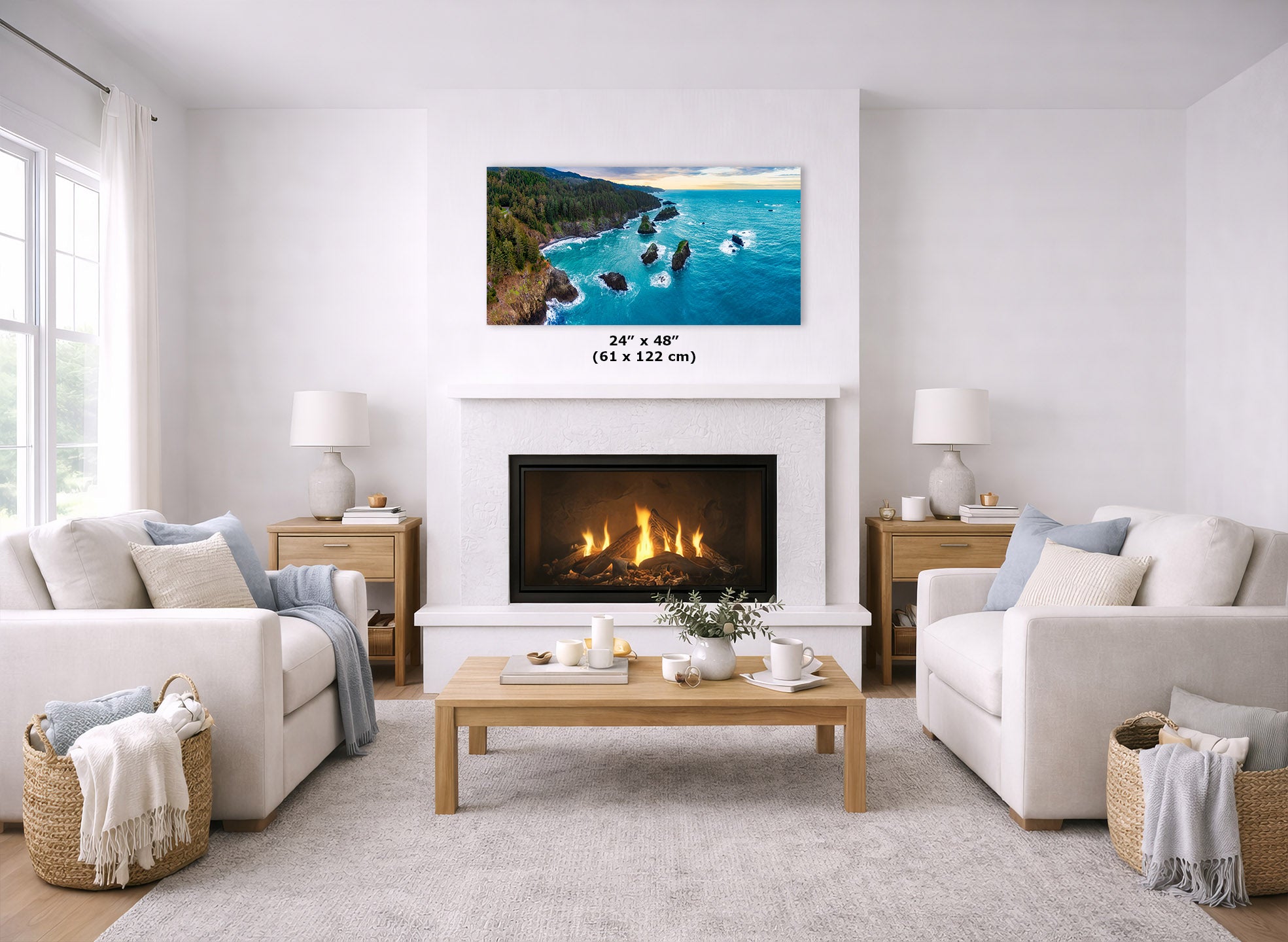 A framed coastal landscape print hangs above a fireplace in a bright, modern living room with white sofas, a wooden coffee table, and baskets. The print shows turquoise water, rocky shoreline, and green hills, with size text reading 24 x 48 (61 x 122 cm) below it.