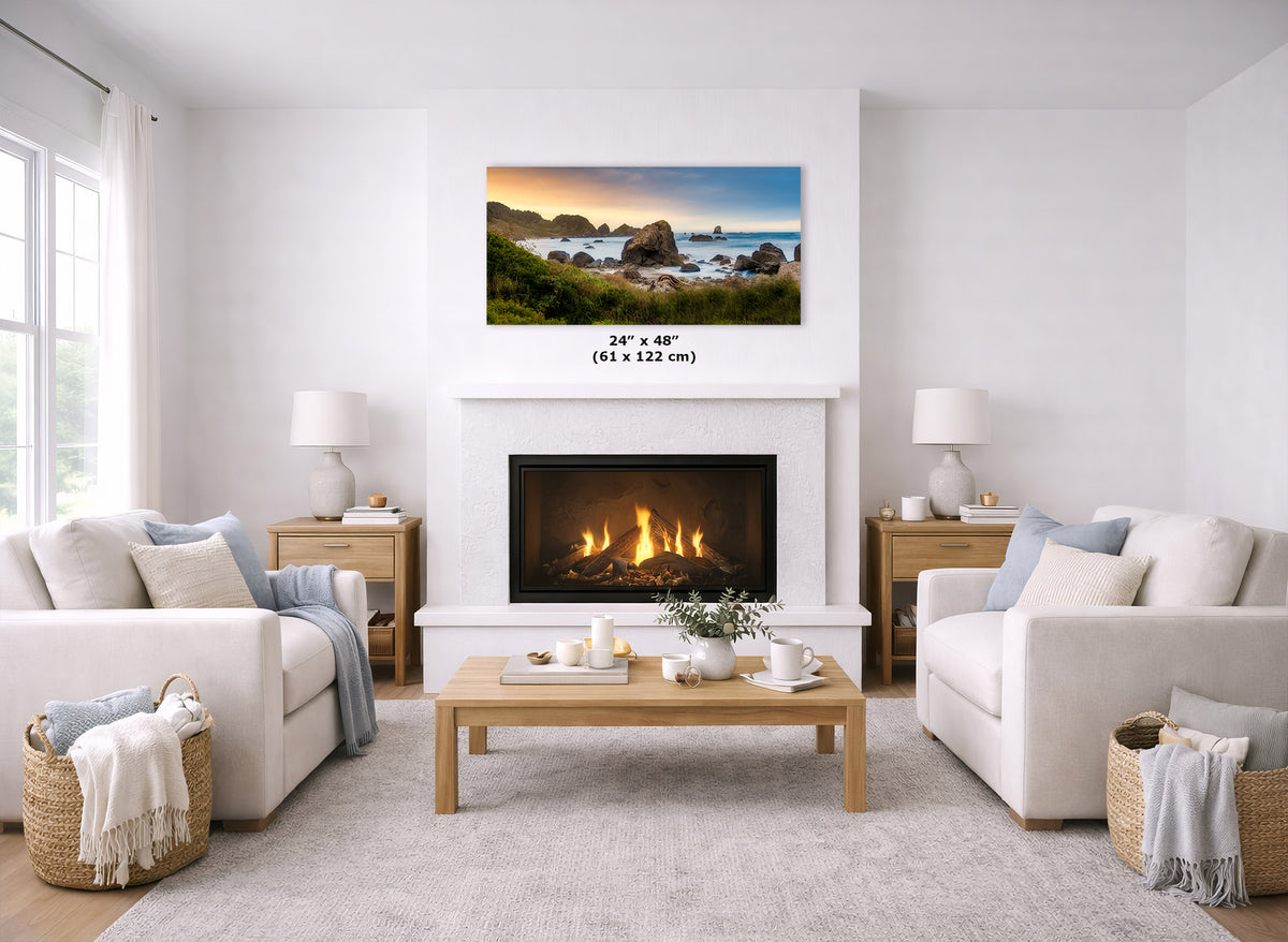 A coastal landscape wall art print is displayed above a fireplace in a bright, modern living room. The print shows a rocky beach and ocean scene with blue water, green cliffs, and a warm sunset sky.