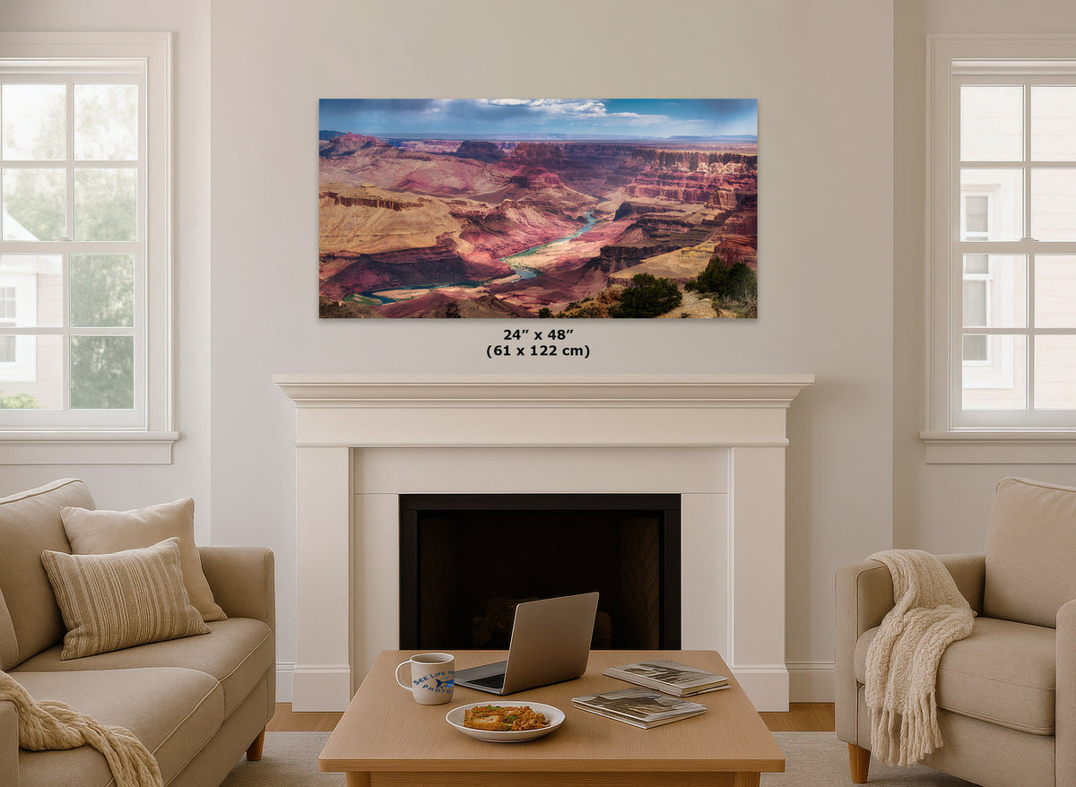 A large rectangular wall art print showing a canyon landscape with a river, displayed above a white fireplace in a bright living room. Small text below the print reads 24" x 48" (61 x 122 cm).