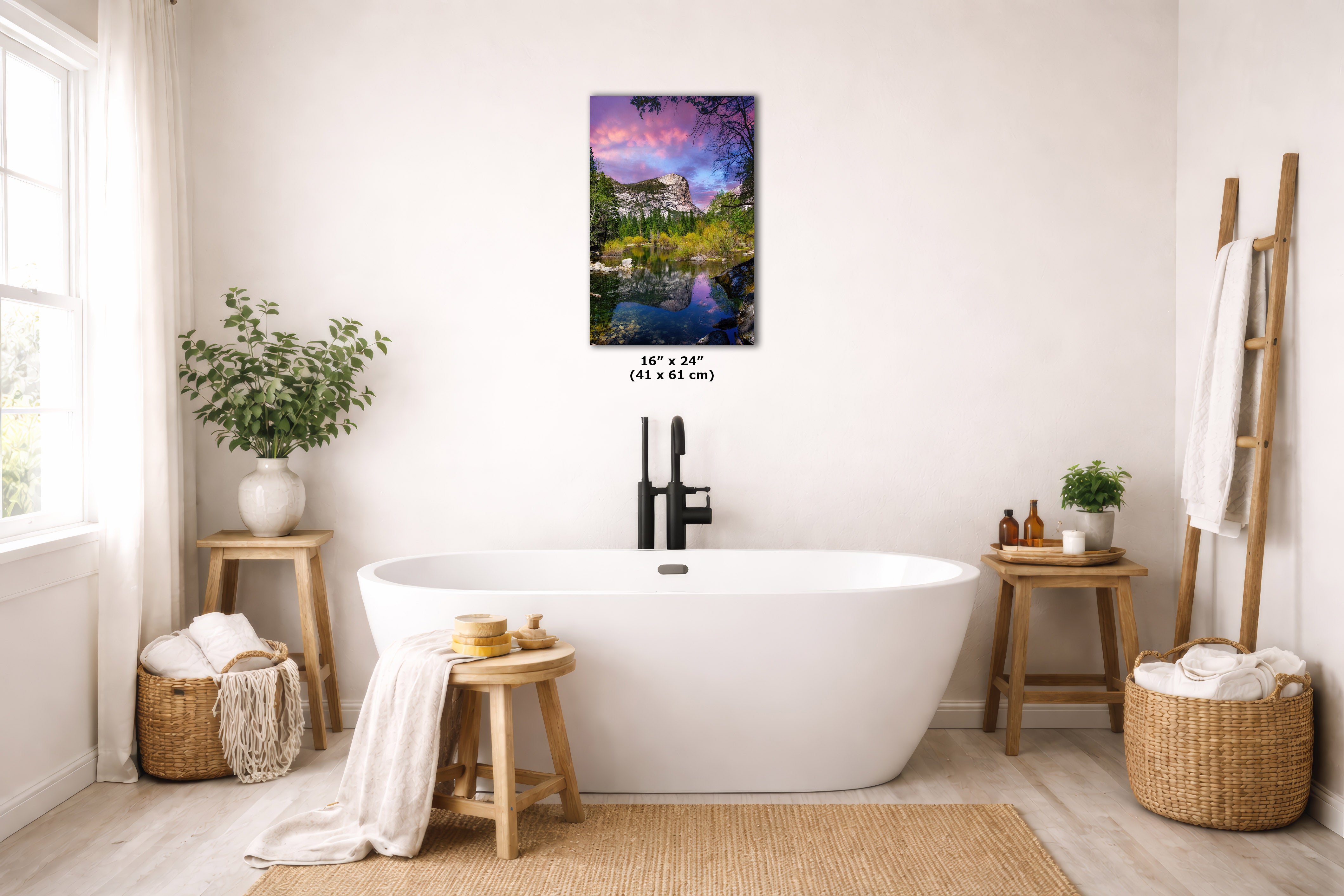 A freestanding white bathtub in a bright, neutral-toned bathroom with a colorful landscape wall art print above it. The print shows a scenic garden pond with a house and a dramatic purple, pink, and blue sky.