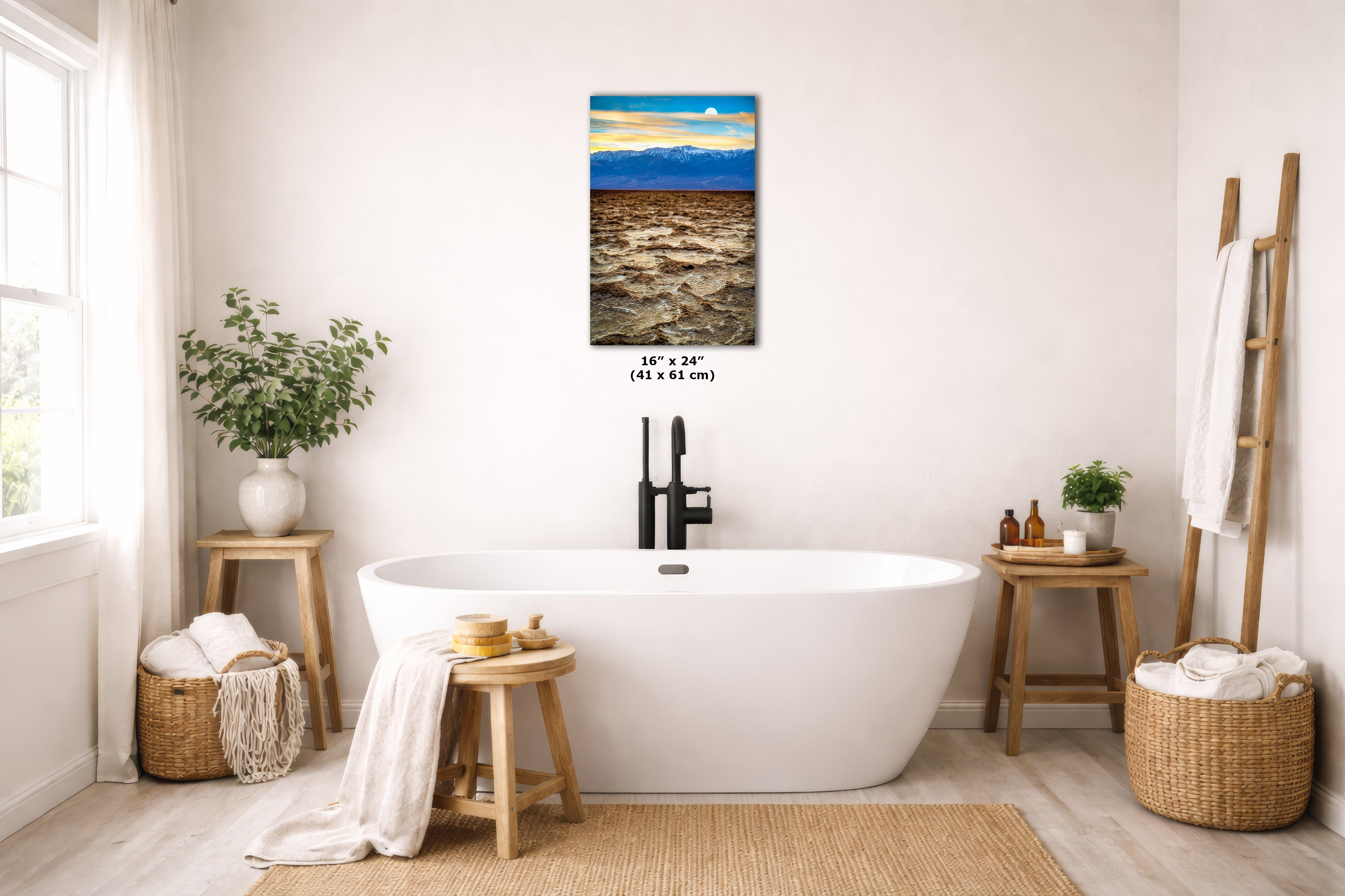 A framed wall art print showing a beach landscape with blue sky and golden sand, displayed above a white freestanding bathtub in a bright, modern bathroom. The print includes size text reading 16 x 24 in (41 x 61 cm).