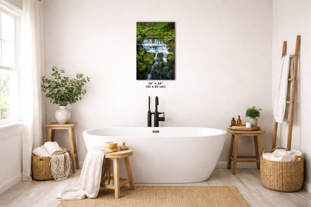 A freestanding white bathtub in a bright, neutral-toned bathroom with a framed wall art print above it showing a waterfall in a lush green forest. The print includes size text reading 16 x 24 inches (41 x 61 cm).