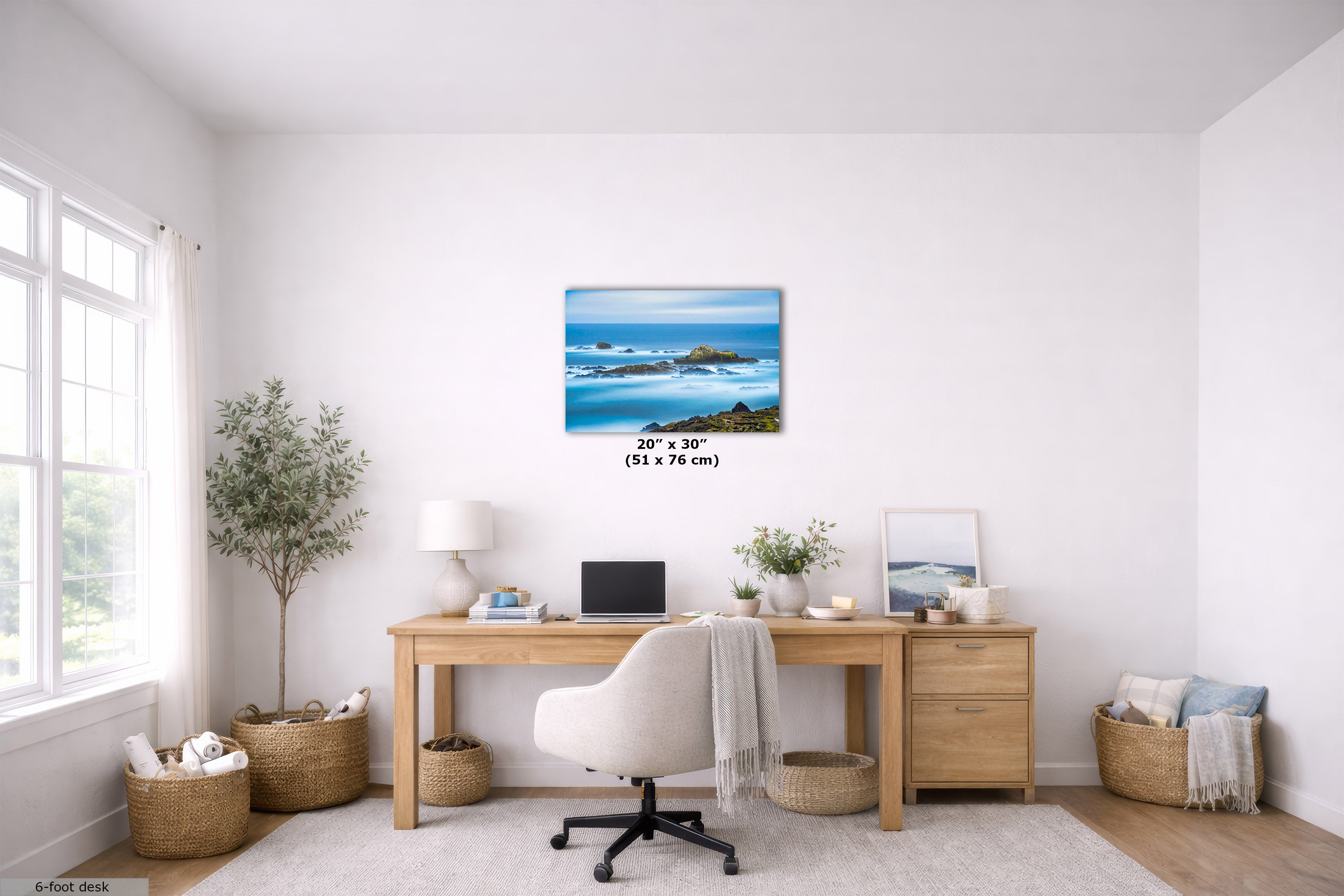 A 23 x 30 inch wall art print depicting a scenic blue ocean and rocky coastline, displayed in a bright, modern home office setting above a wooden desk.